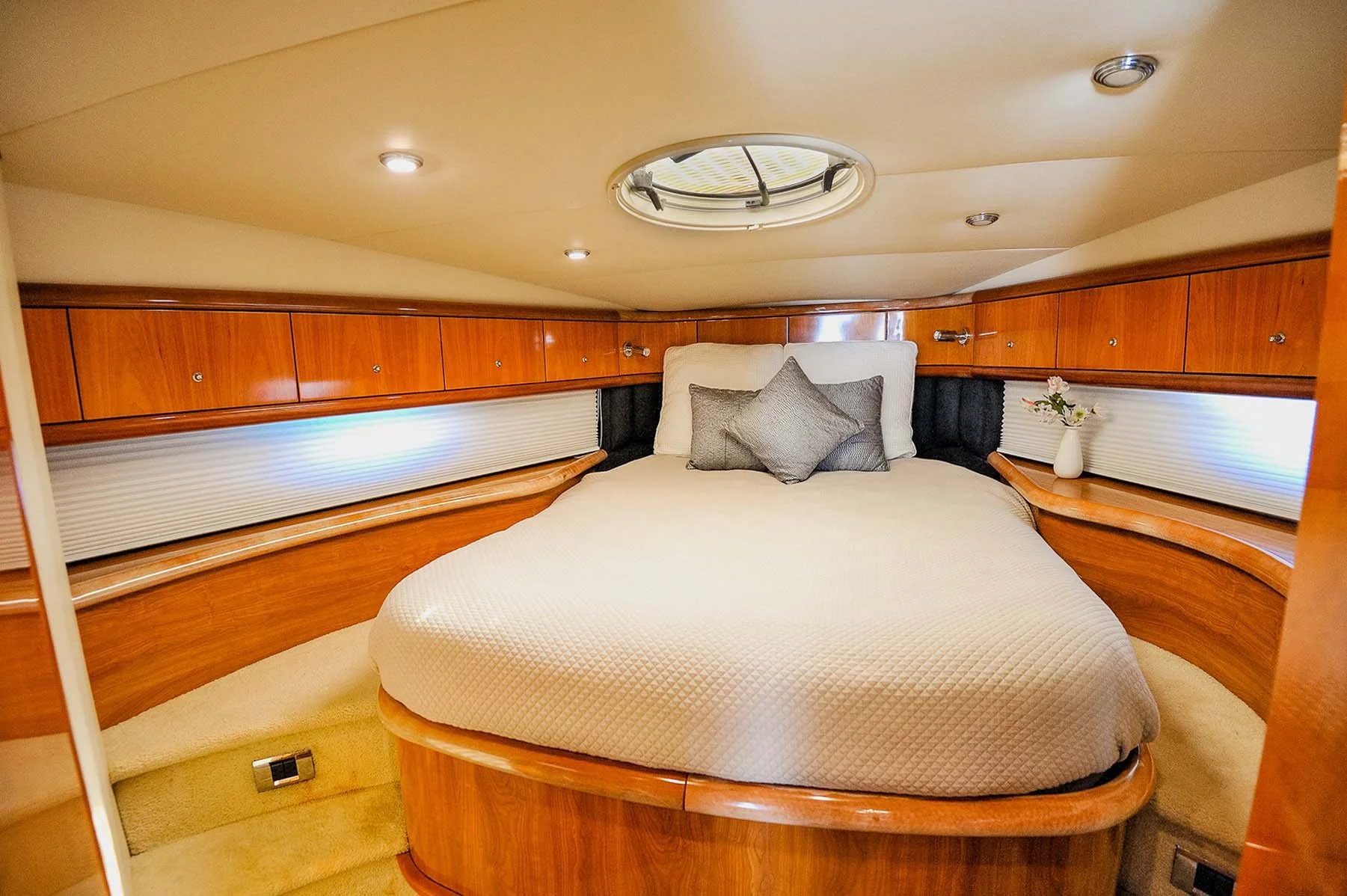 A cozy bedroom with a circular bed, gray pillows, wooden cabinetry above the bed, a small vase with flowers to the side, and a porthole window in the ceiling.