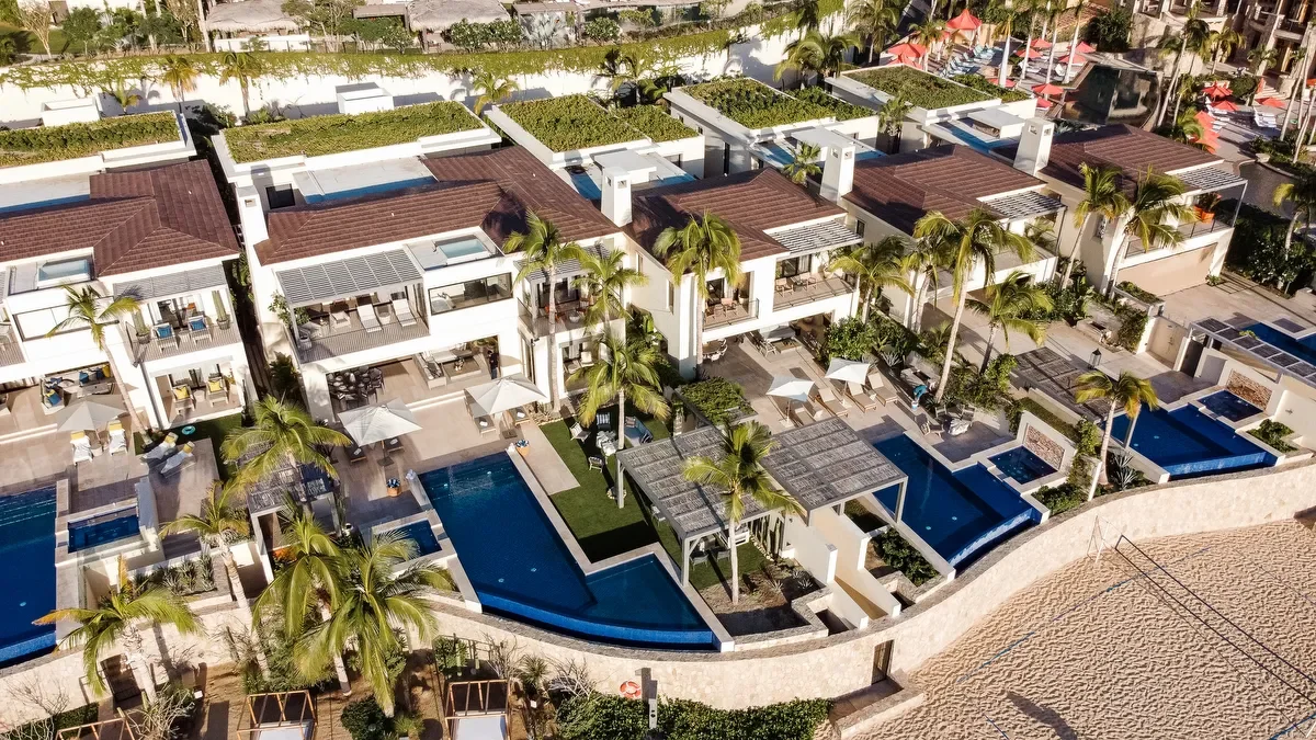 Aerial view of a luxury resort with multiple modern villas featuring private pools and tropical landscaping, including palm trees.