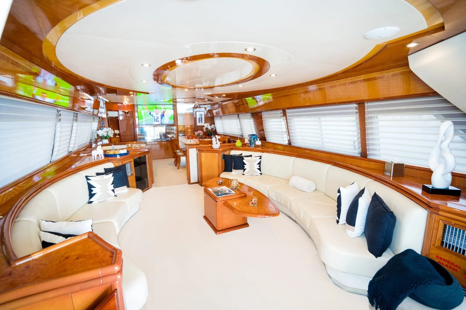 Luxurious yacht interior with cream-colored sofa, black and white pillows, wooden accents, large windows with white blinds, and a flat-screen TV on the wall.