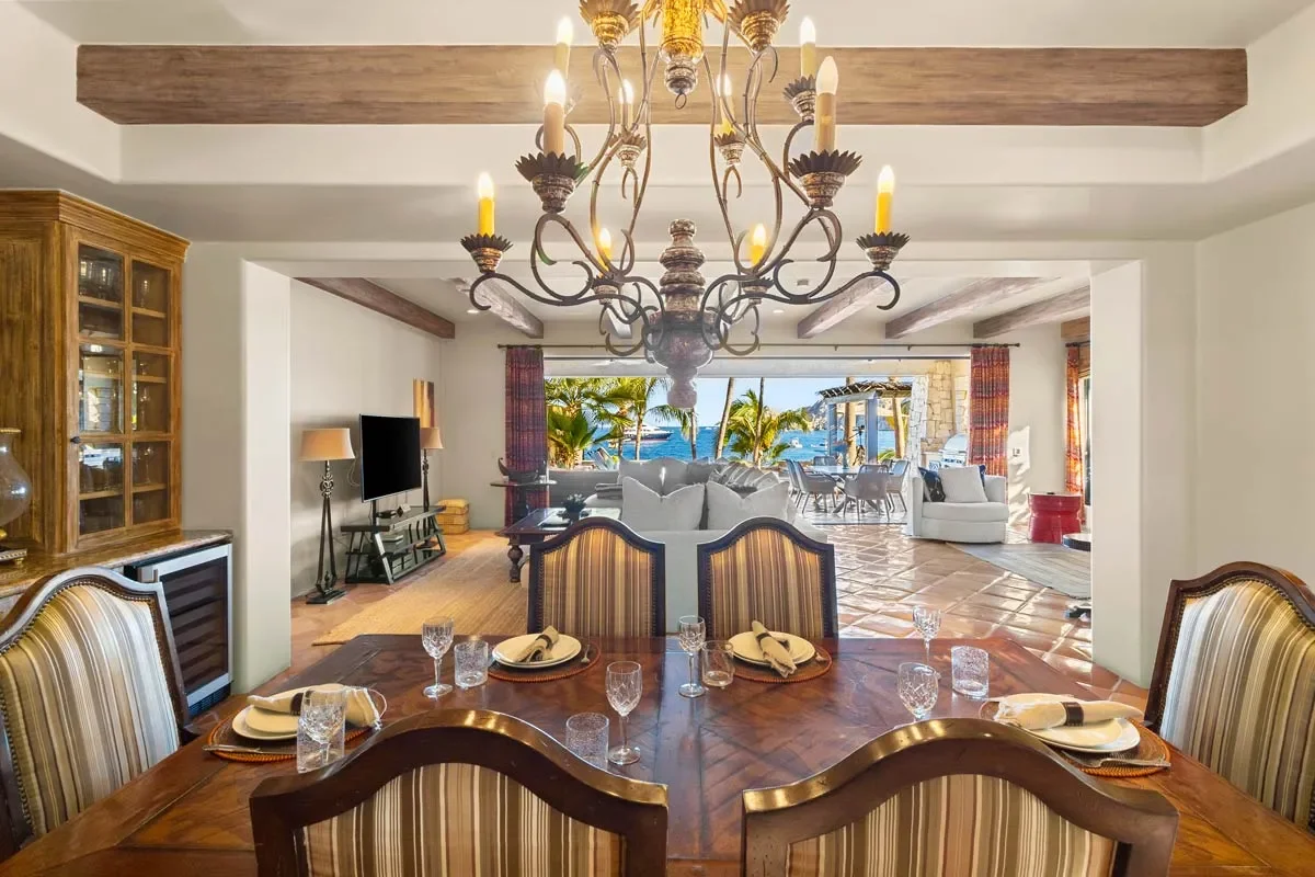 Dining room with a large wooden table set with plates, glasses, and napkins, surrounded by striped upholstered chairs, chandelier overhead, open view to living room and outdoor patio with palm trees and ocean in the background.