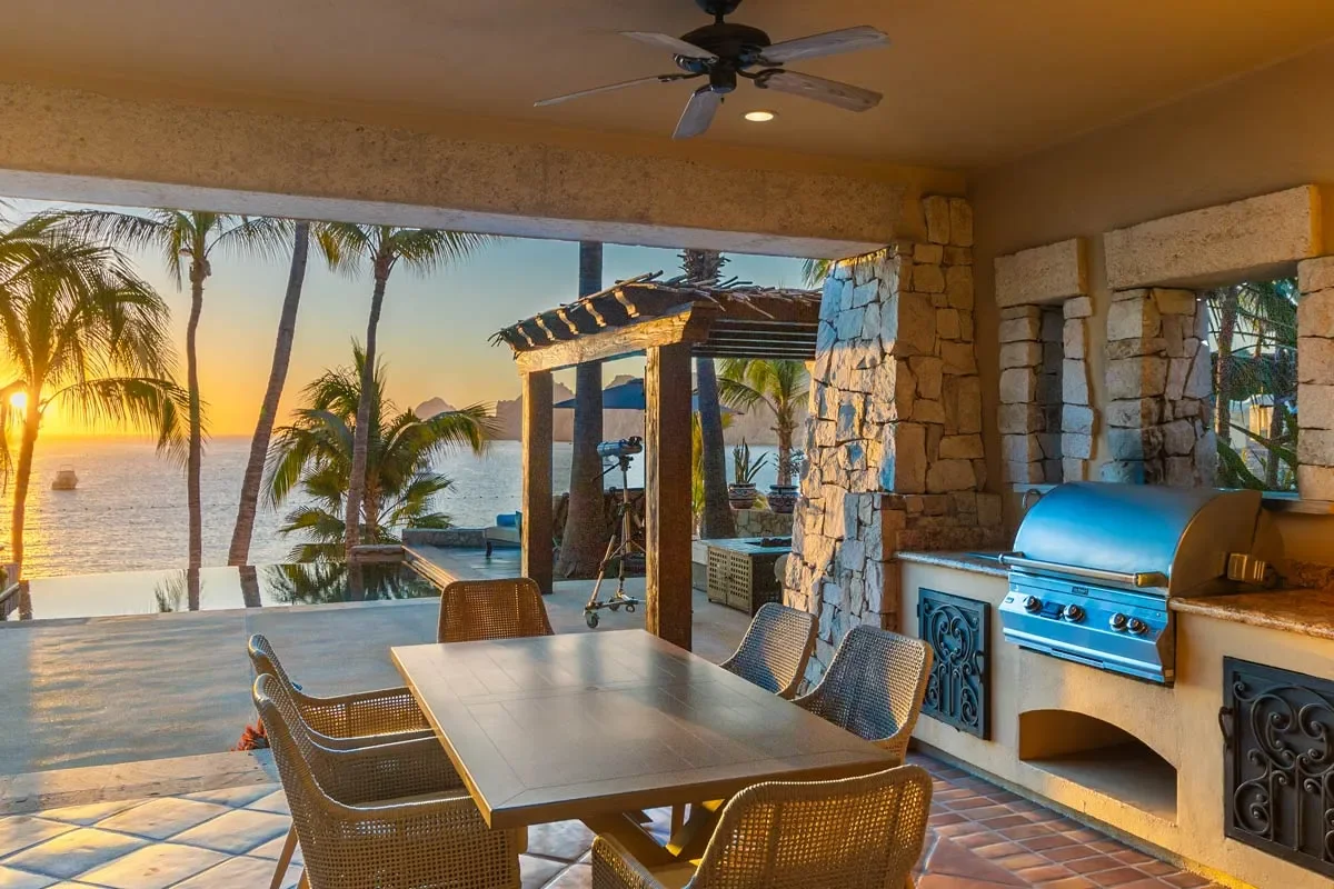 Outdoor patio with a dining table and chairs, stone grill, and telescope, overlooking a tropical sunset with palm trees and the ocean in the background.