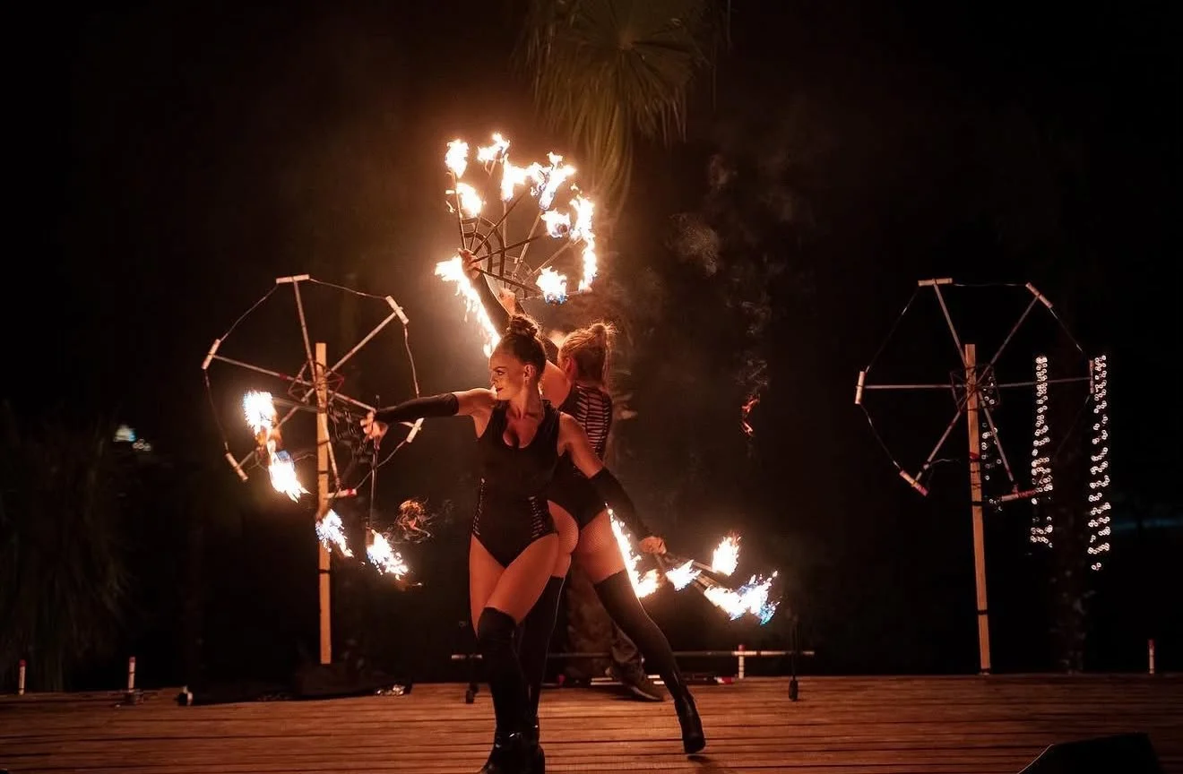 Two female fire performers dancing with flaming fire tools on an outdoor stage at night, with large fire props in the background and palm trees behind them.