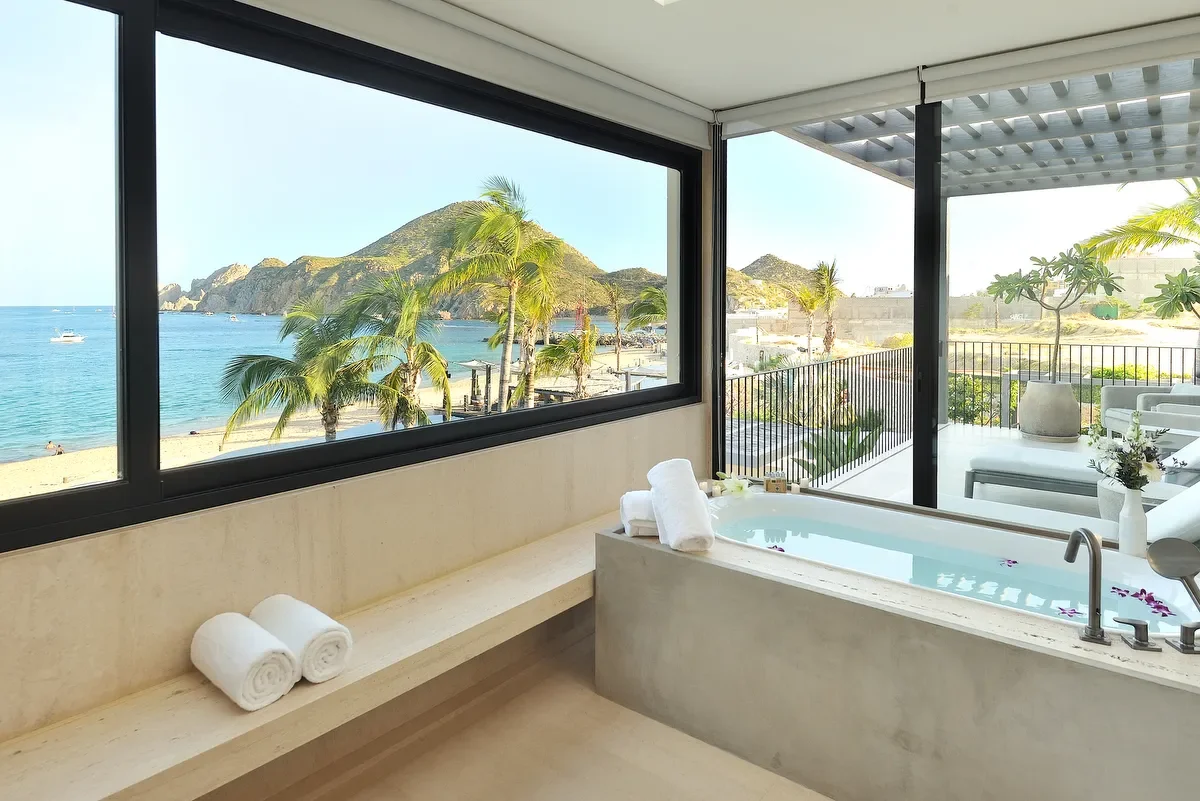 Indoor bathroom with a bathtub overlooking a beach with palm trees, mountains, and ocean, seen through large windows and sliding glass doors.
