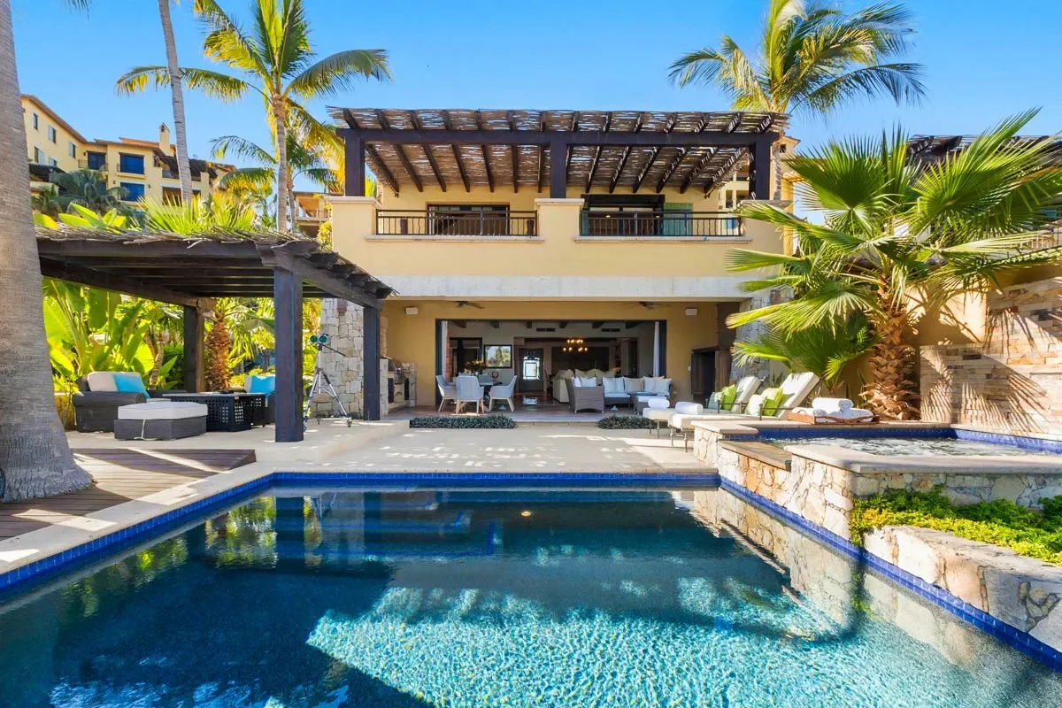 Luxury villa with a swimming pool, outdoor seating, palm trees, and a two-story house with a balcony under a clear blue sky.
