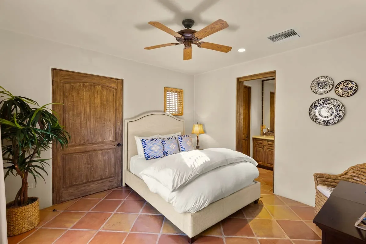 A bedroom with a beige bed, blue patterned pillows, a nightstand with a yellow lamp, a ceiling fan, a plant in a woven basket, decorative plates on the wall, and wooden door and closet frames.