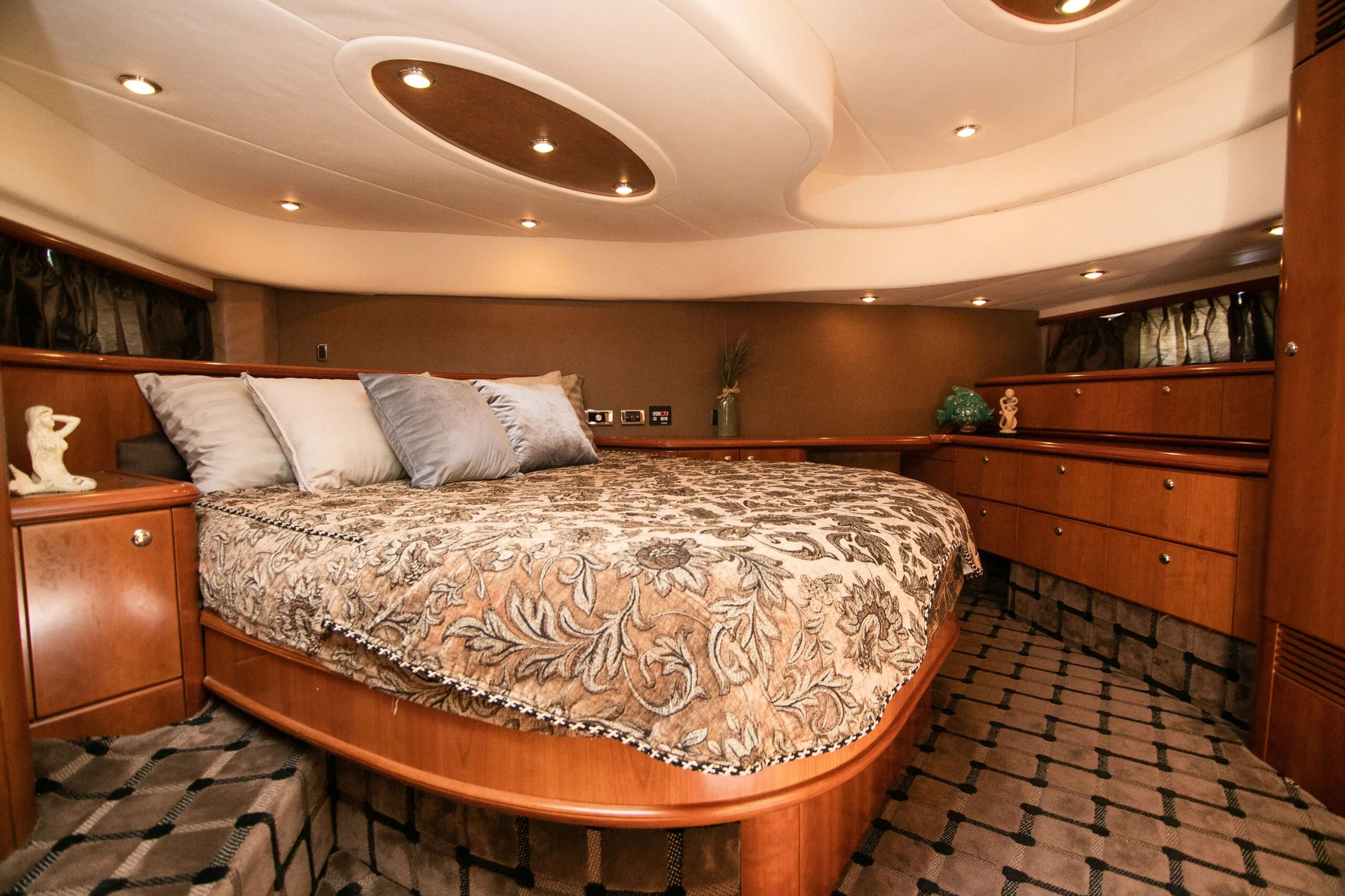 A cozy bedroom with a bed covered in a patterned beige and brown bedspread and multiple pillows. The room features wooden built-in drawers and floating shelves, with various decorations such as small sculptures and a vase with a plant. The ceiling ha