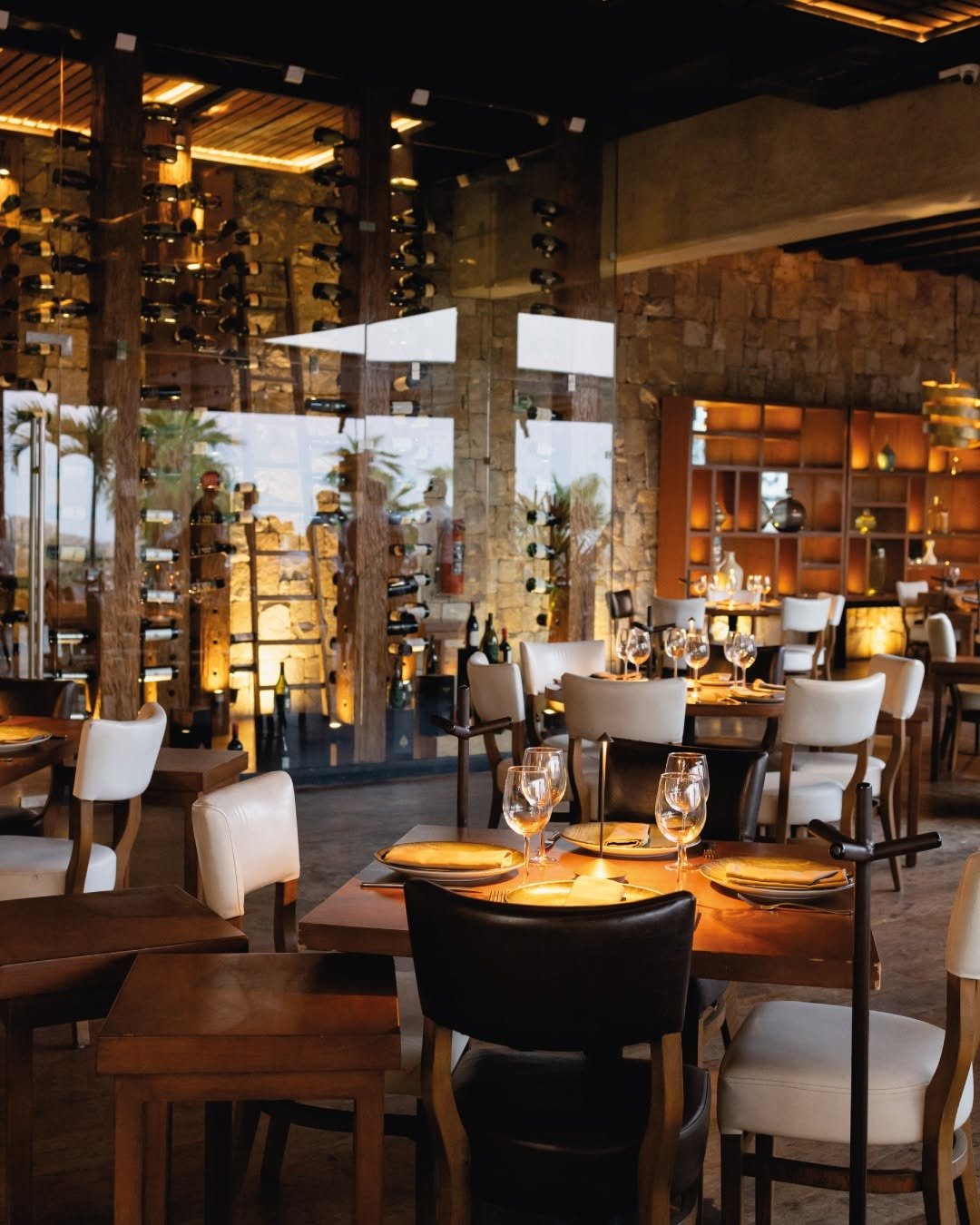 A stylish restaurant with wooden tables and white and black chairs, set with wine glasses, napkins, and plates. The background features a wine rack, a stone wall, and warm lighting, creating an inviting ambiance.
