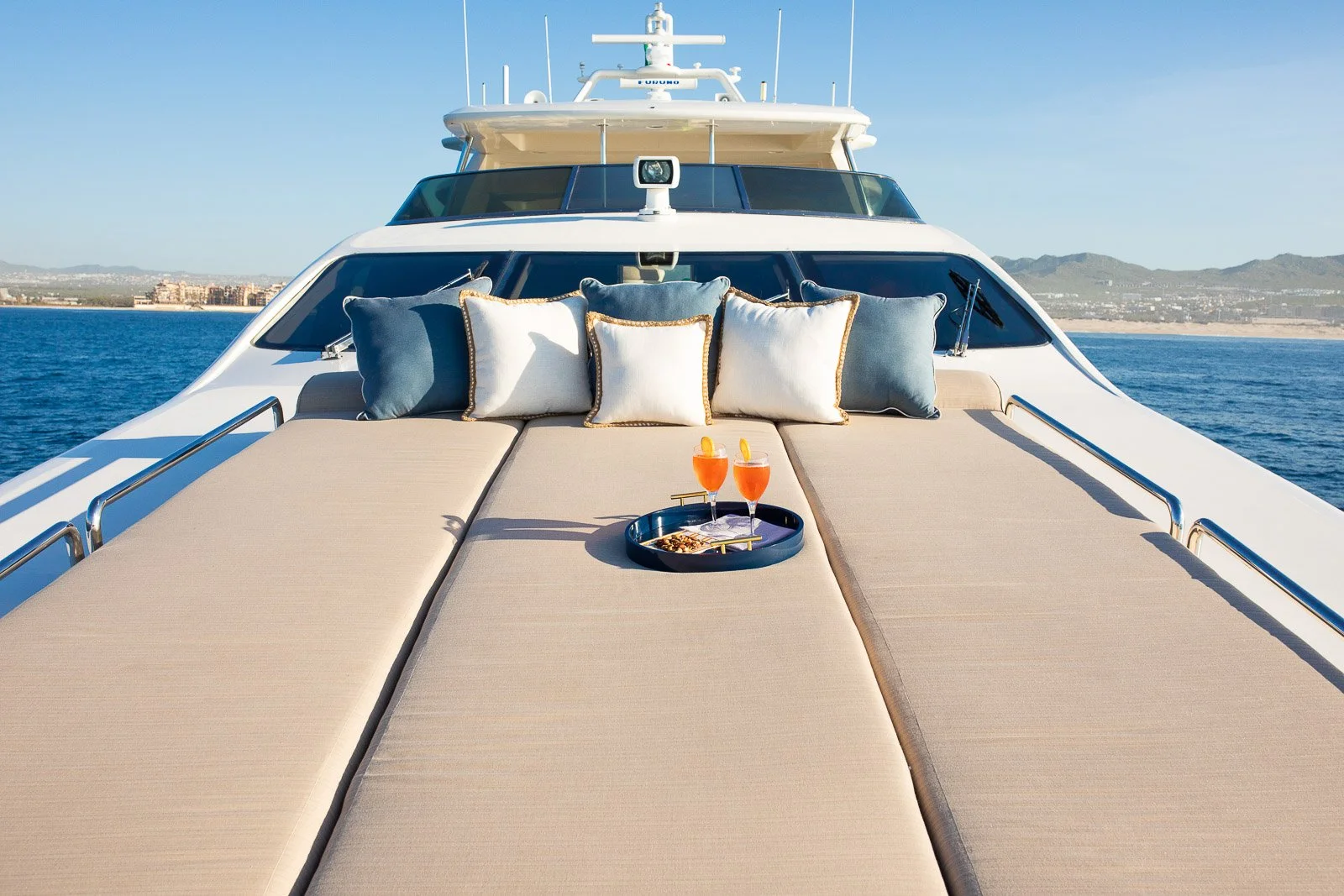 The deck of a yacht with beige sunbeds, several pillows in shades of blue, white, and beige, two drinks with lemon slices on a tray, and a view of the ocean and distant landforms.