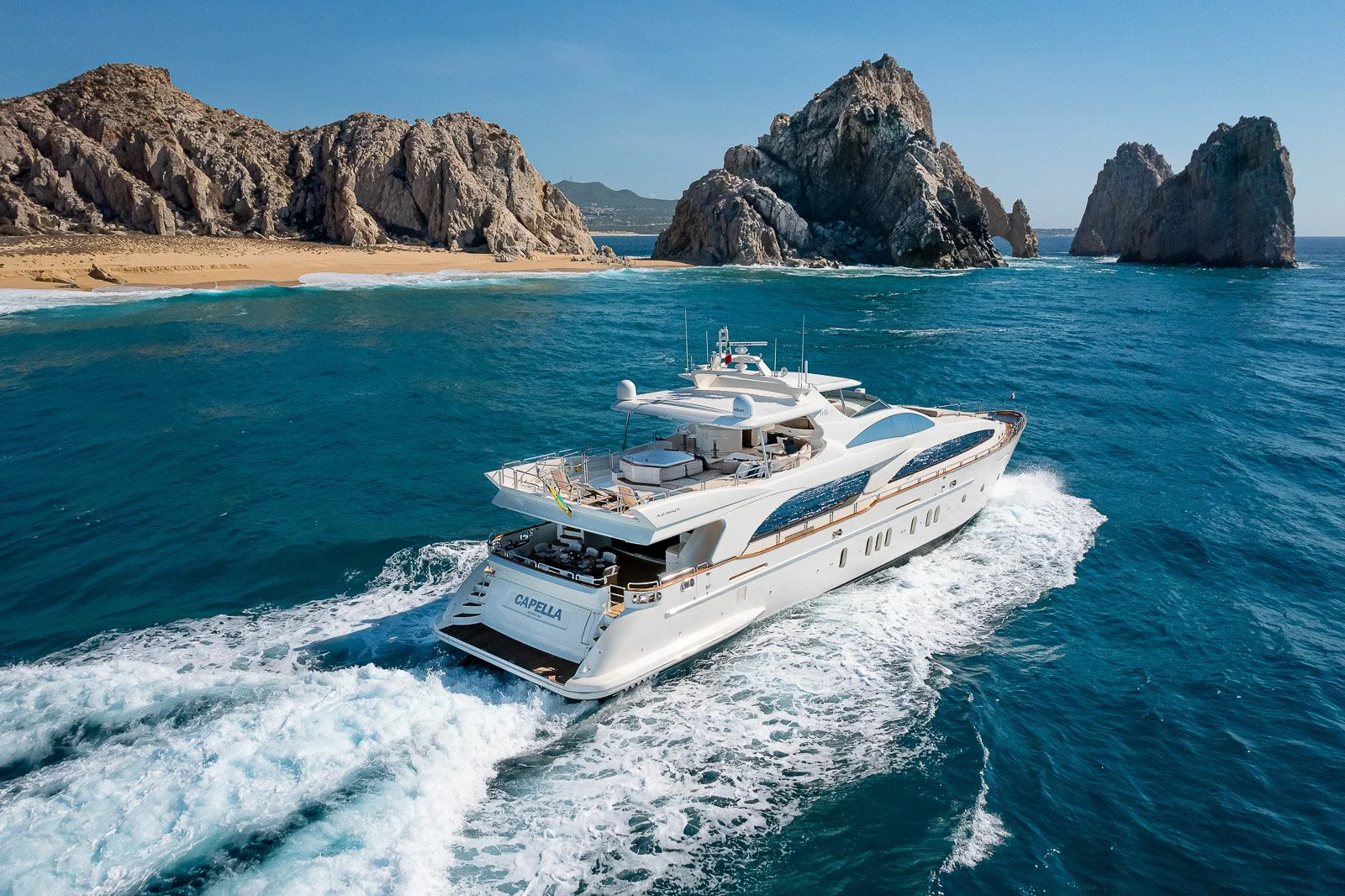 A luxury white yacht sailing in blue ocean waters near rocky cliffs and sandy beach coastline.