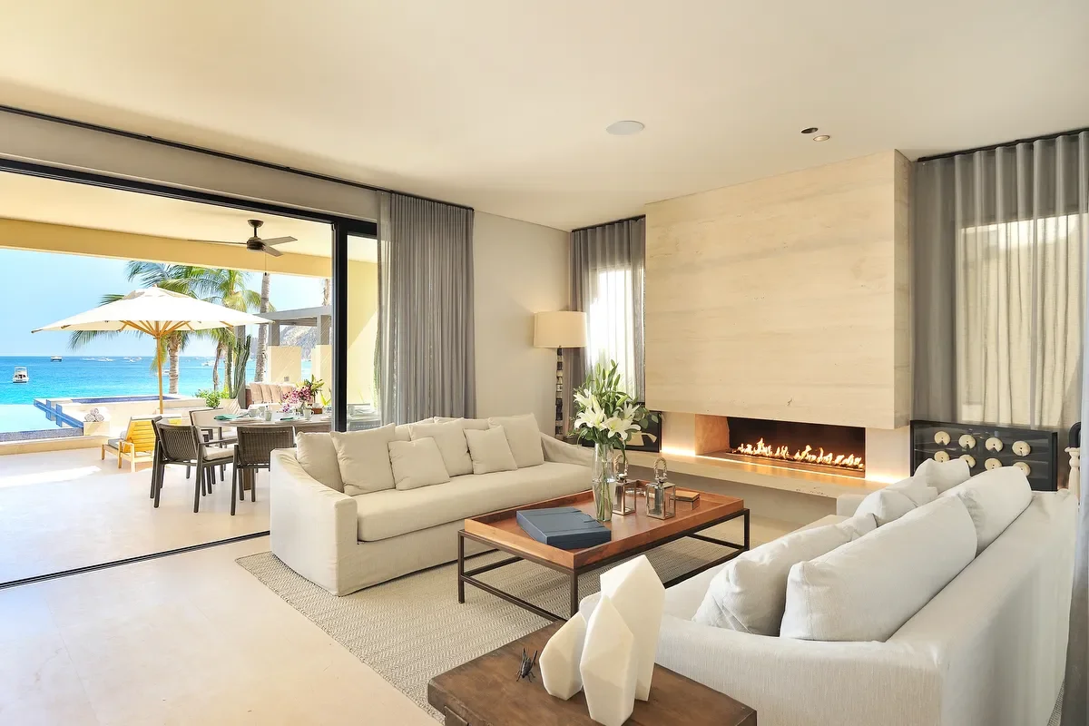 Living room with white sofas, a fireplace, large windows with curtains, and a view of a patio with outdoor dining and a pool overlooking the ocean with palm trees.