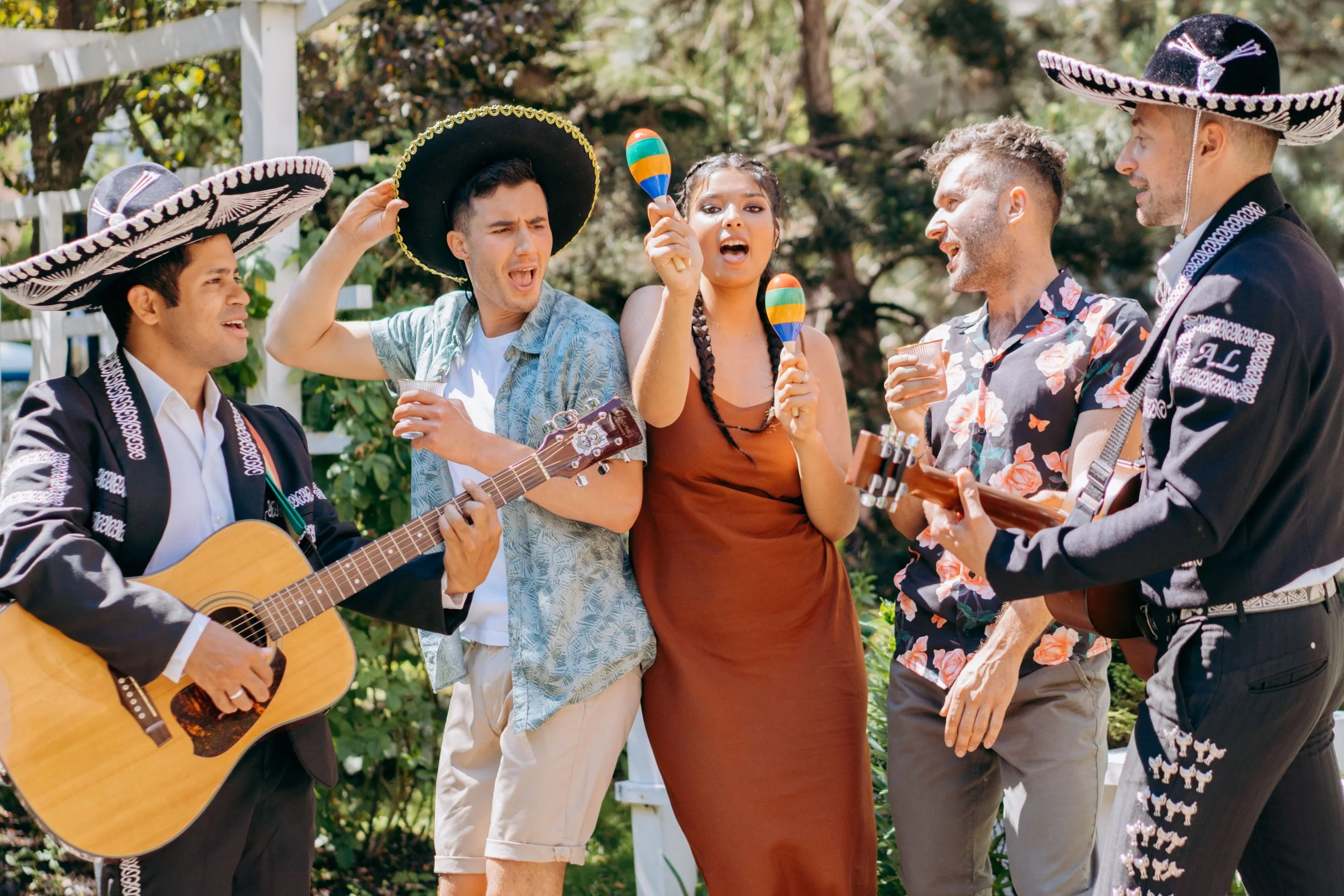 Group of people at an outdoor celebration wearing traditional Mexican sombreros, singing, playing guitar, and holding maracas.