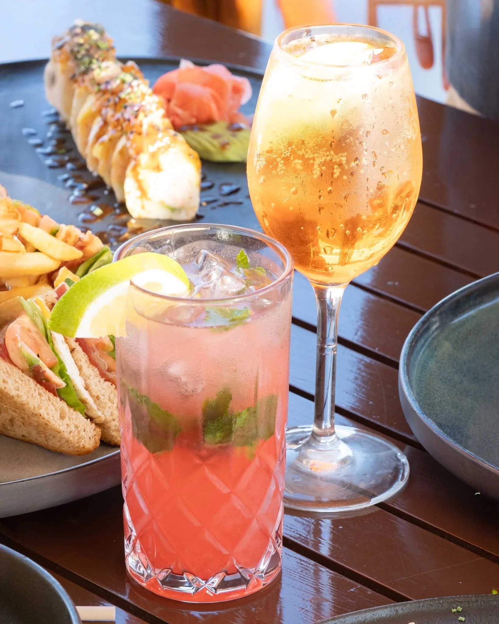 A pink-colored cocktail drink with lime wedge garnish in a patterned glass, set on a wooden table. In the background are sushi rolls with ginger slices and a dark plate with sauce. A soda or similar beverage in a clear glass with ice and mint leaves is in front of the cocktail.