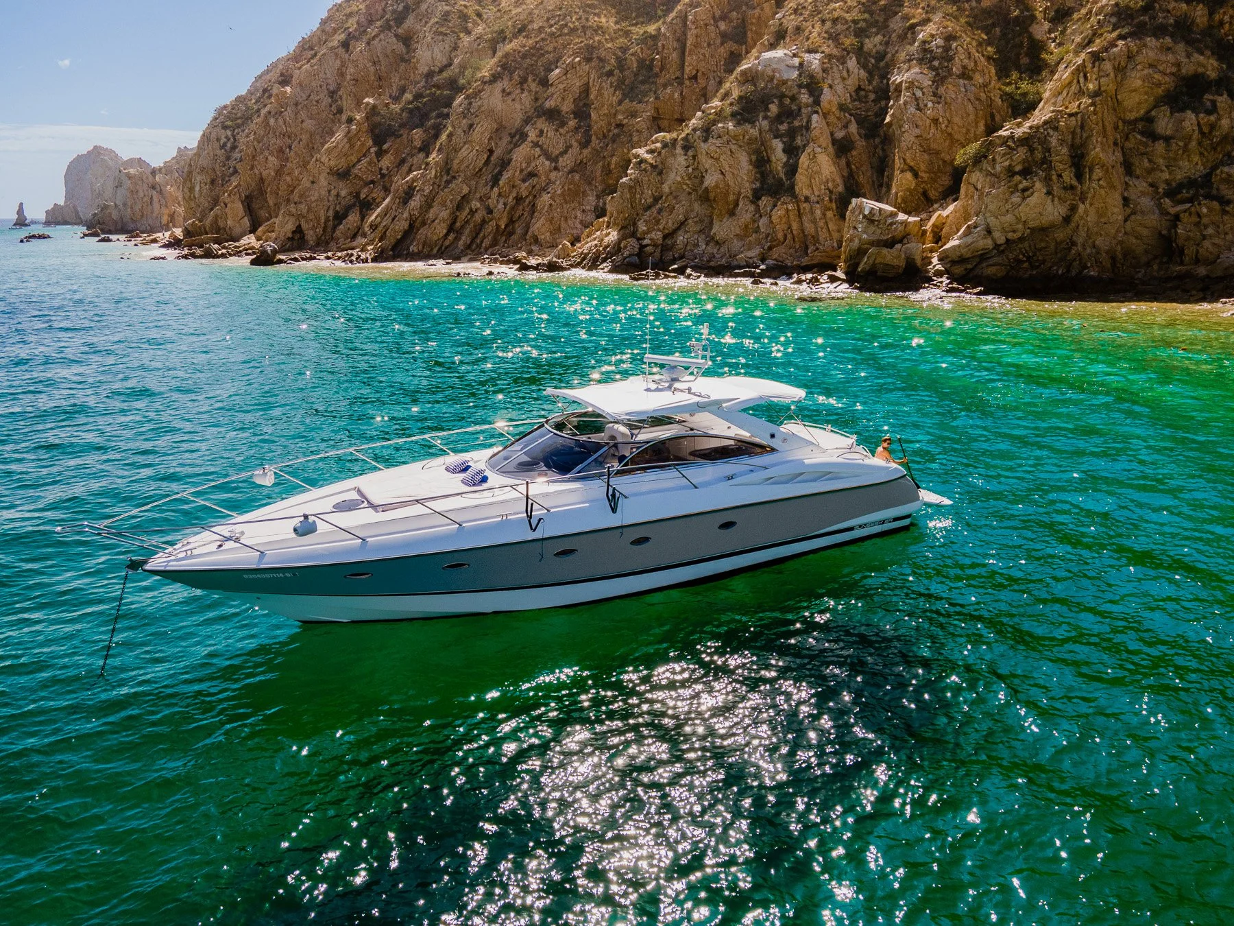 A white yacht floating near a rocky coastline with turquoise water and sunlight reflecting on the surface.