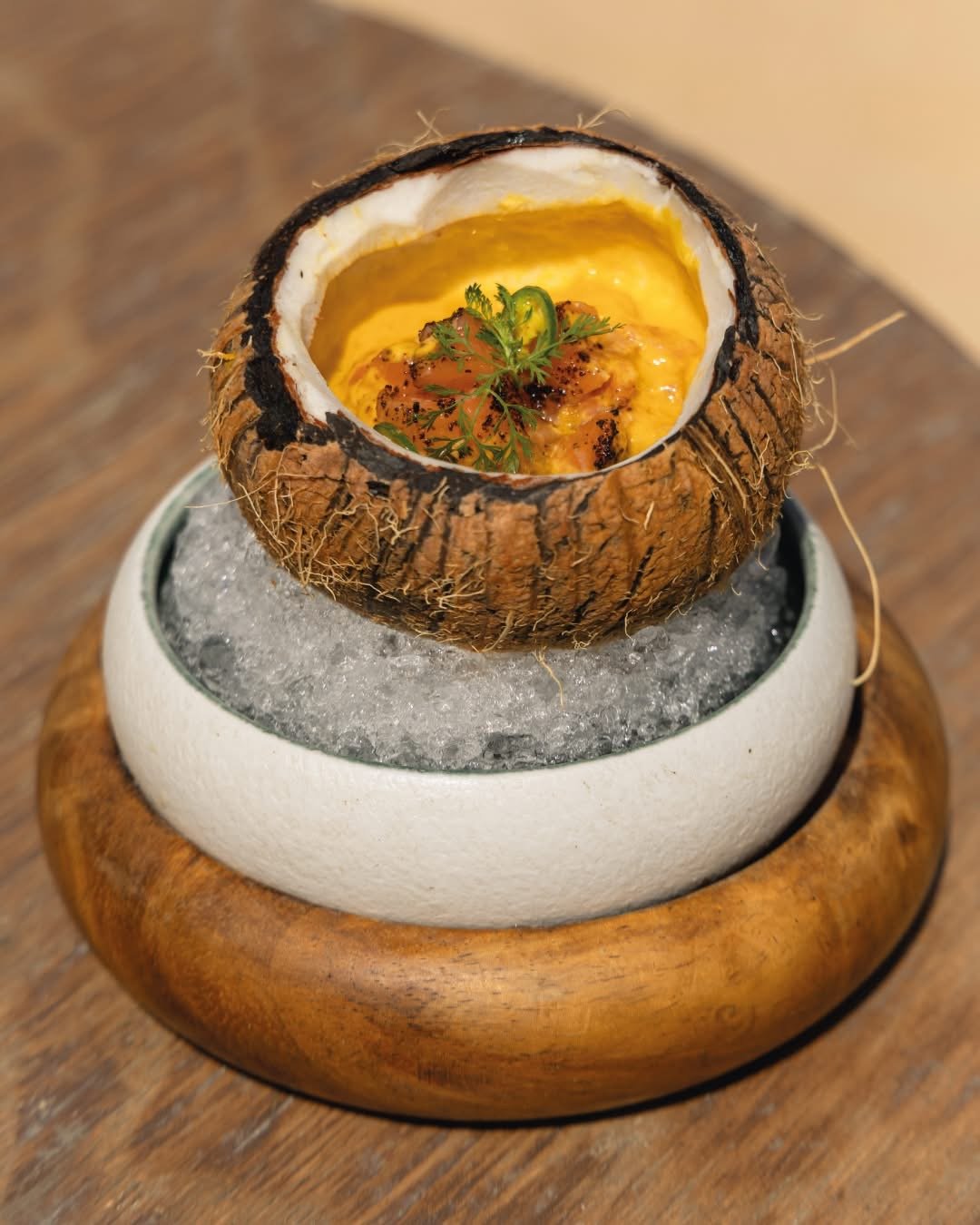 Baked dish served inside a hollowed-out coconut shell, garnished with herbs, placed on a bed of ice in a ceramic bowl on a wooden table.