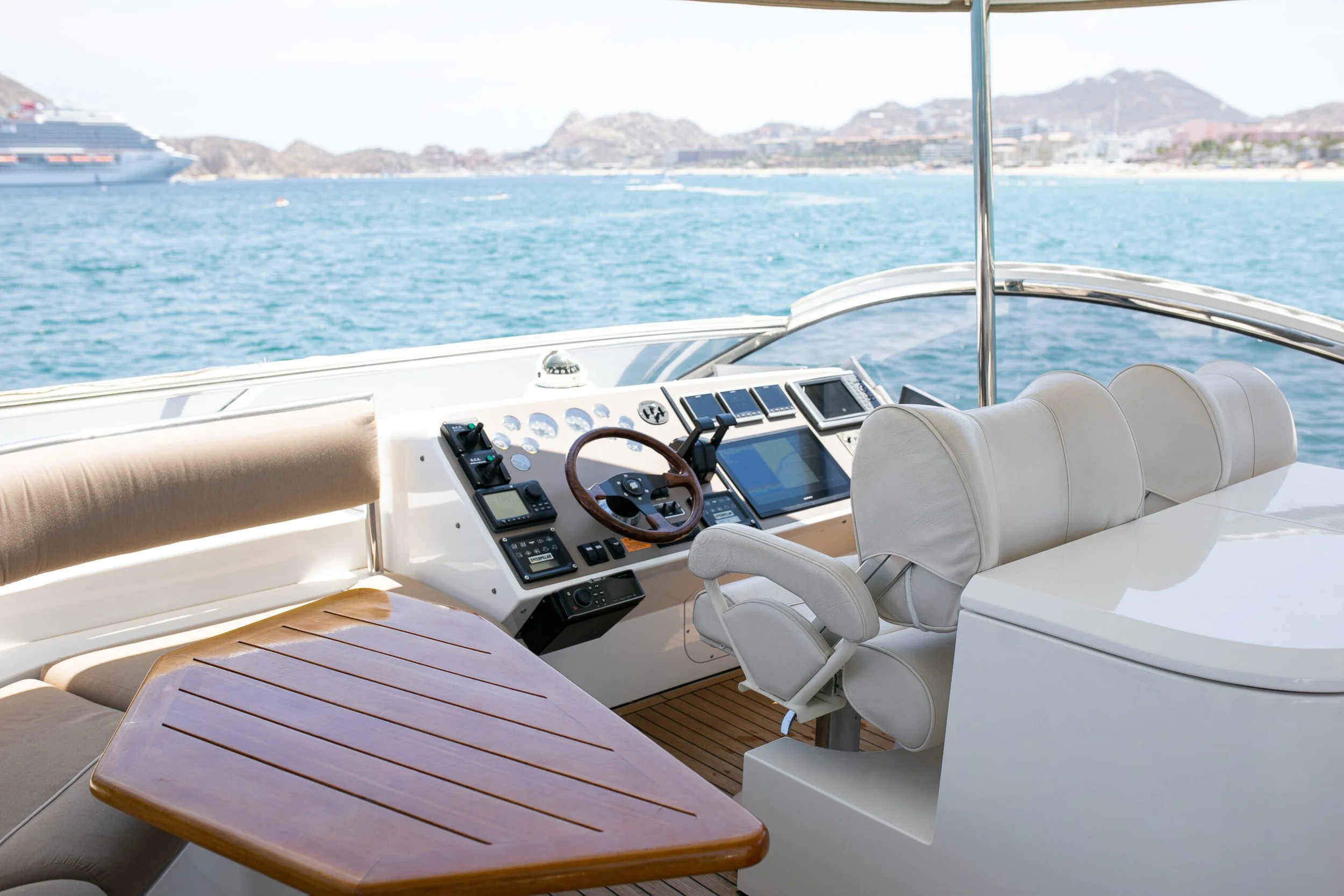 View from the deck of a yacht showing a control console with navigation equipment, a beige cushioned seat, a wooden table, and a cityscape with hills in the background across the water.