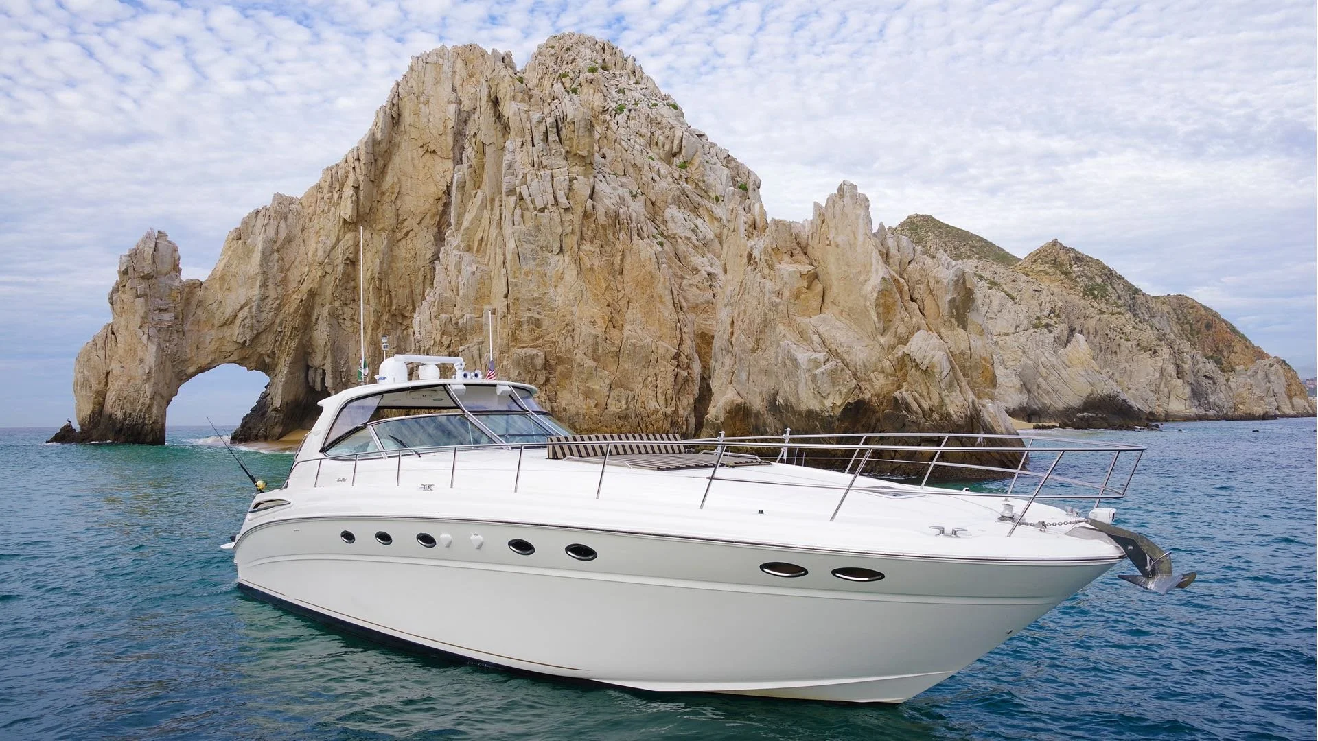 A white yacht floating near a large rock formation with a natural arch on the water, under a partly cloudy sky.