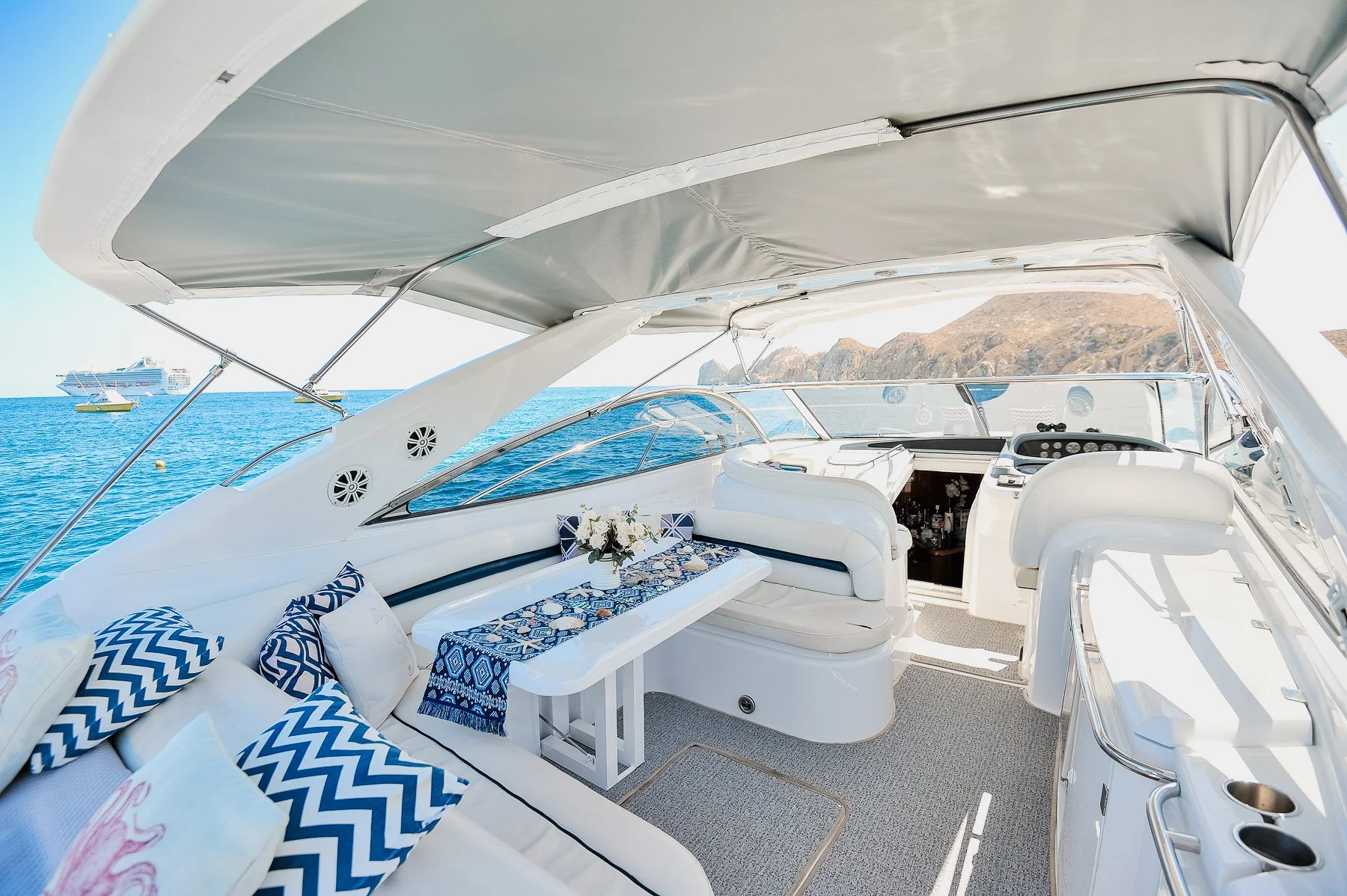 Luxurious yacht deck with seating area, table with a blue patterned tablecloth and flowers, view of the sea with boats and mountains in the background.