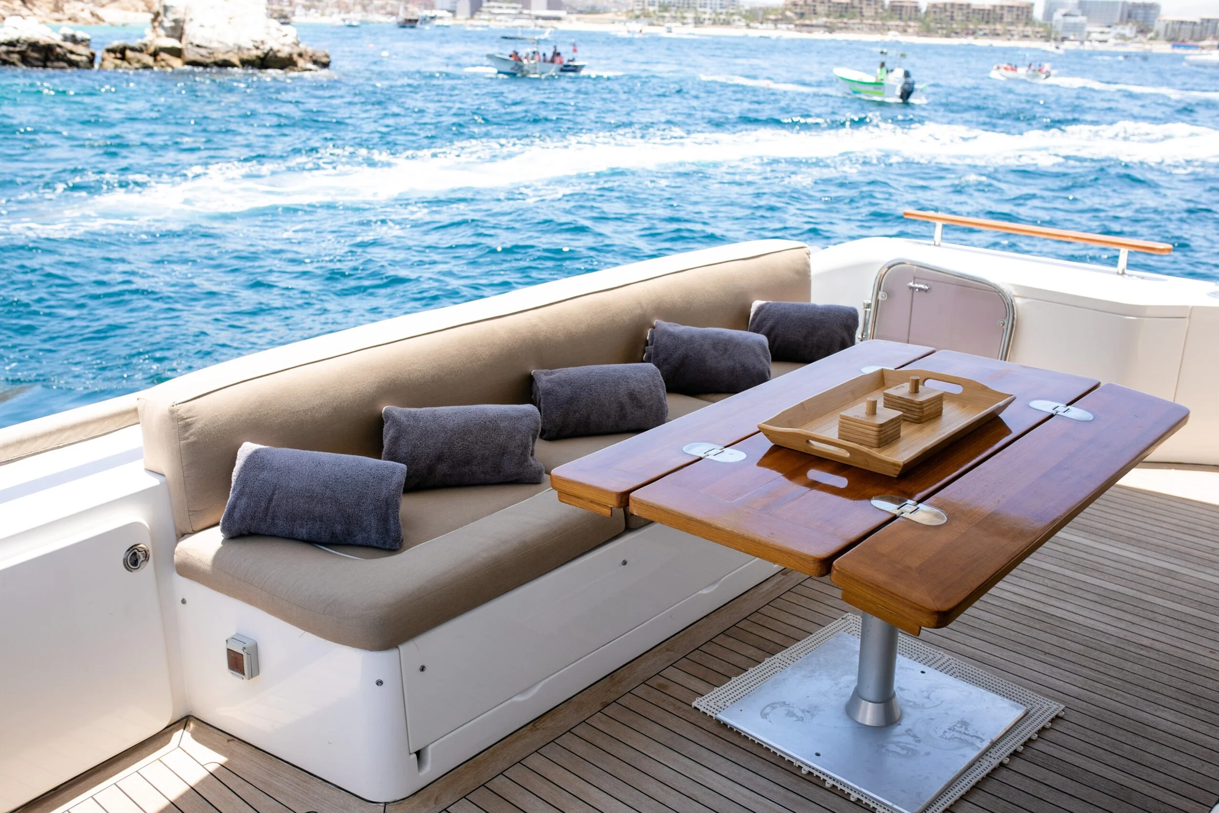 View from a boat showing a seating area with beige and gray cushions and a wooden table, with water, boats, and a city skyline in the background.