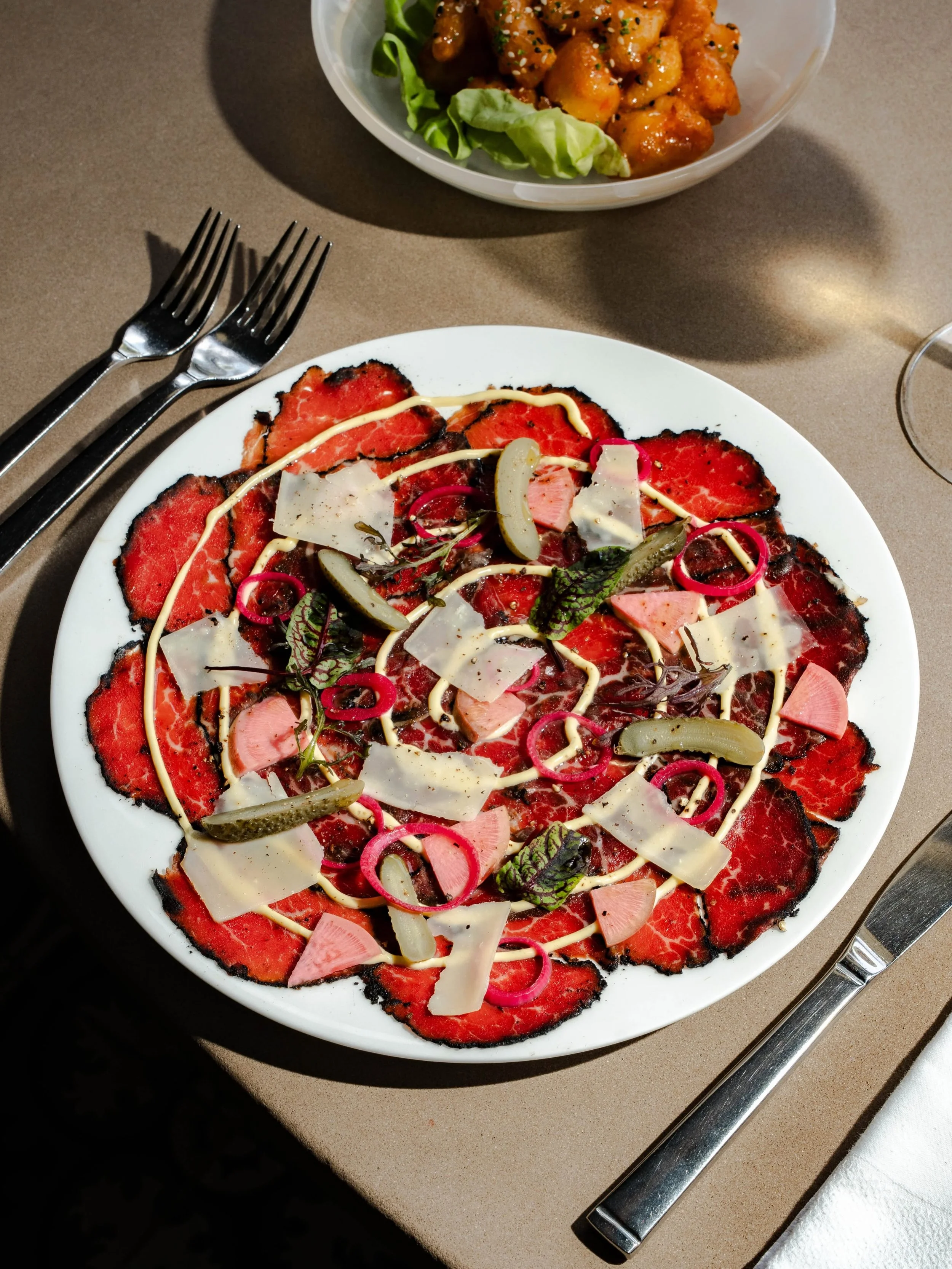 A white plate with a thin crust pizza topped with red meat, shredded cheese, and pickled vegetables, with two forks placed on the left side and a knife on the right side. In the background, there is a bowl of fried chicken pieces garnished with sesame seeds and herbs, resting on a piece of lettuce.