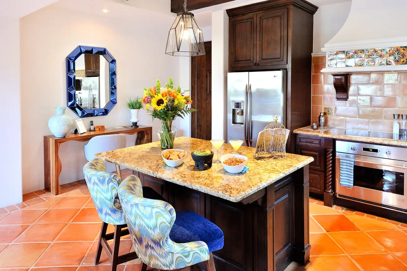 A kitchen with terracotta tile flooring, dark wood cabinetry, a granite island with food and drinks, a vase of colorful flowers, a decorative mirror, and modern appliances including a stainless steel refrigerator and oven.