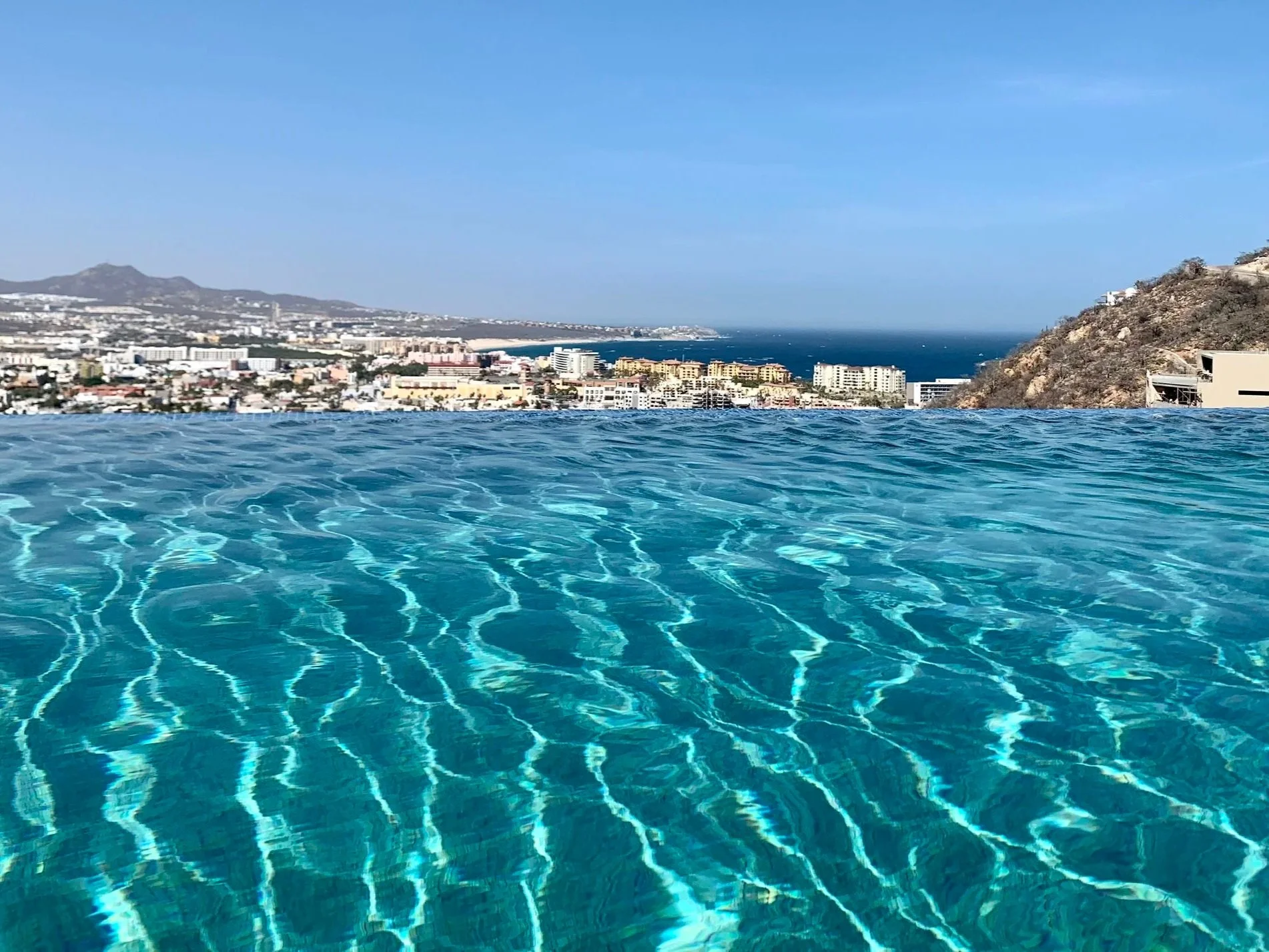 View of a city skyline with buildings, mountains in the background, and an ocean or large body of water in the distance, taken from a location with a swimming pool in the foreground.