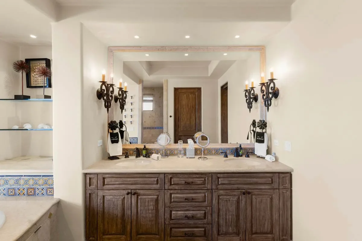 Bathroom vanity with a large mirror, dark wood cabinets, two black wall-mounted candle-style lights, and various toiletries on the countertop, with a glimpse of a shower area in the background.