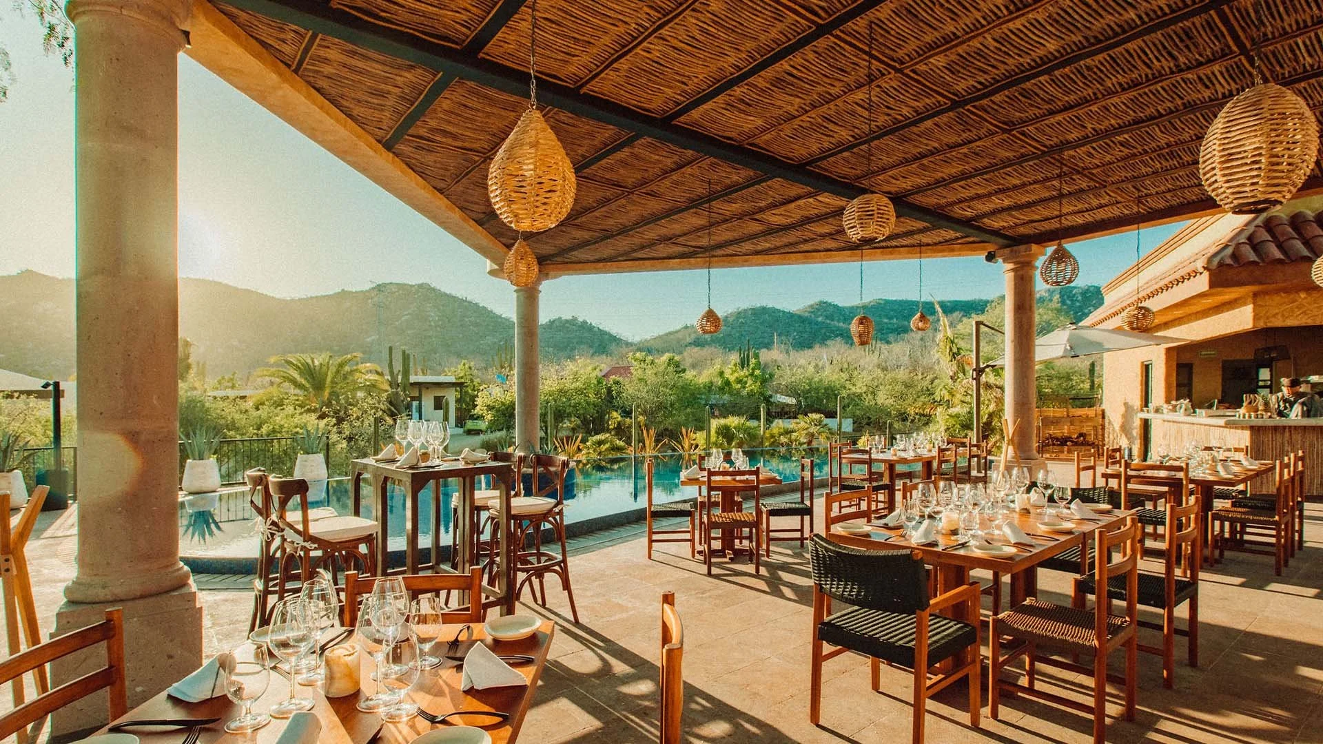 Outdoor restaurant with wooden tables and chairs, set with glasses and white napkins, under a thatched roof with hanging wicker lanterns, overlooking a scenic landscape with mountains and greenery.