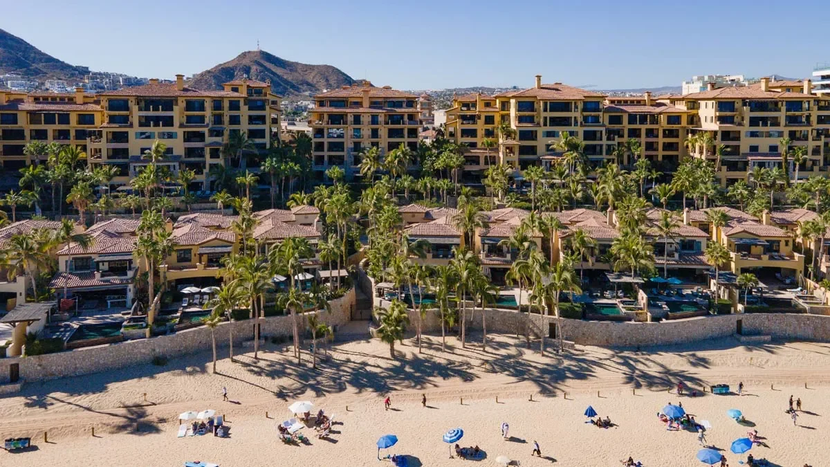 Aerial view of beachside resort with multi-story buildings, palm trees, and people on the sandy beach with umbrellas.