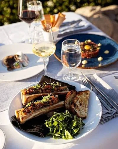 A table set for outdoor dining with plates of food, glasses of white and red wine, and a napkin with cutlery.