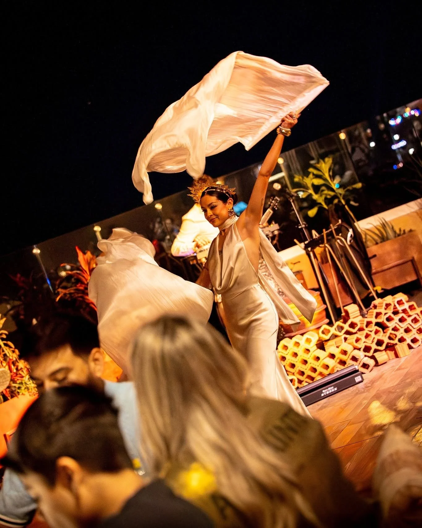 A woman dressed in white, wearing a crown and jewelry, performs a dance with flowing fabric in front of an audience at a nighttime event, with warm lighting and decorative elements in the background.