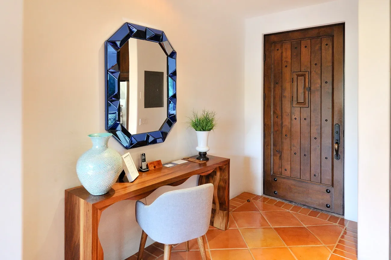 Interior entryway with a wooden door, marble floor tiles, a wooden console table with decorative items including a large vase, a mirror with a blue beveled frame, and a small chair with a pillow.