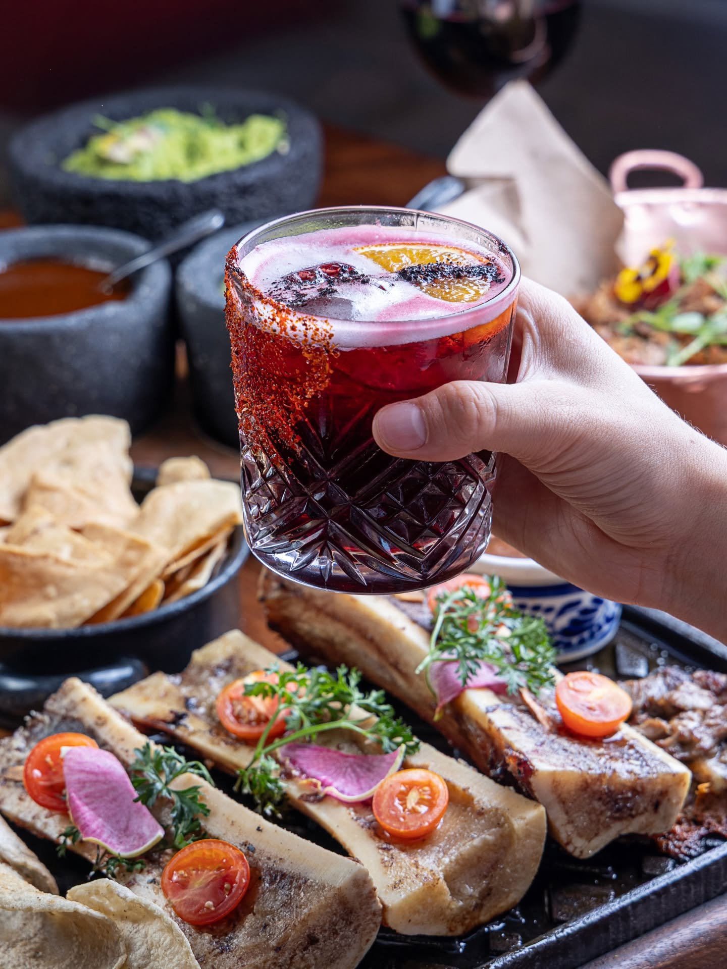 Hand holding a glass of red drink with citrus slices, surrounded by Mexican food including quesadillas, tacos, chips, and salsa.