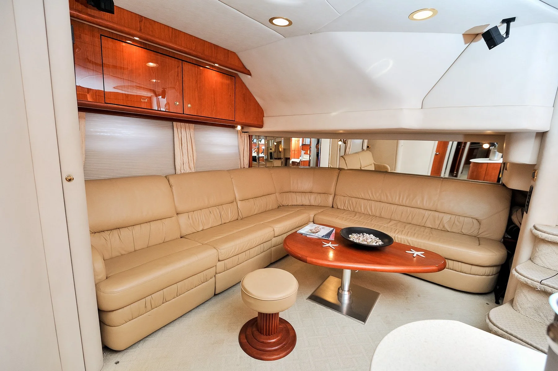 A beige L-shaped leather sectional sofa with a small round beige ottoman and a wooden coffee table with seashells and starfish on it, in a room with wood-paneled and mirrored walls.