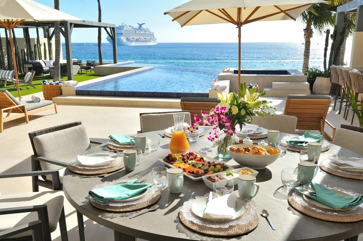 Outdoor dining table set for breakfast on a terrace overlooking the ocean, with a cruise ship in the distance, surrounded by umbrellas and palm trees.