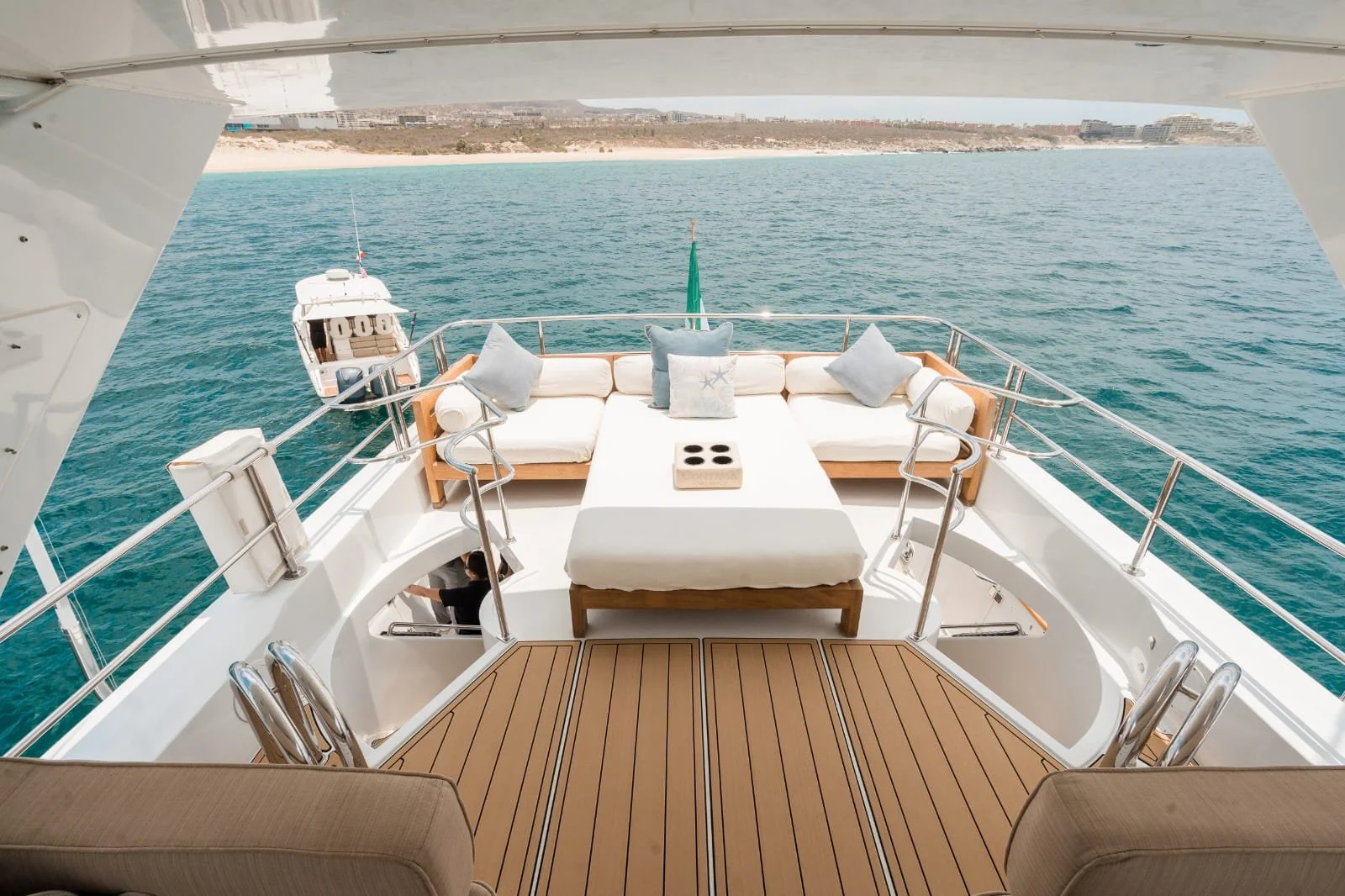 View from a yacht showing a seating area with white cushions and pillows, a small table with a sectional and a cushion, overlooking the blue ocean with another boat in the distance, off a shoreline with buildings.