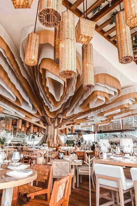 A restaurant with a large, artistic ceiling installation resembling a tree with layered, fabric-like leaves, and hanging woven basket lights. The dining area features wooden tables and chairs, with many diners and waiter staff present.