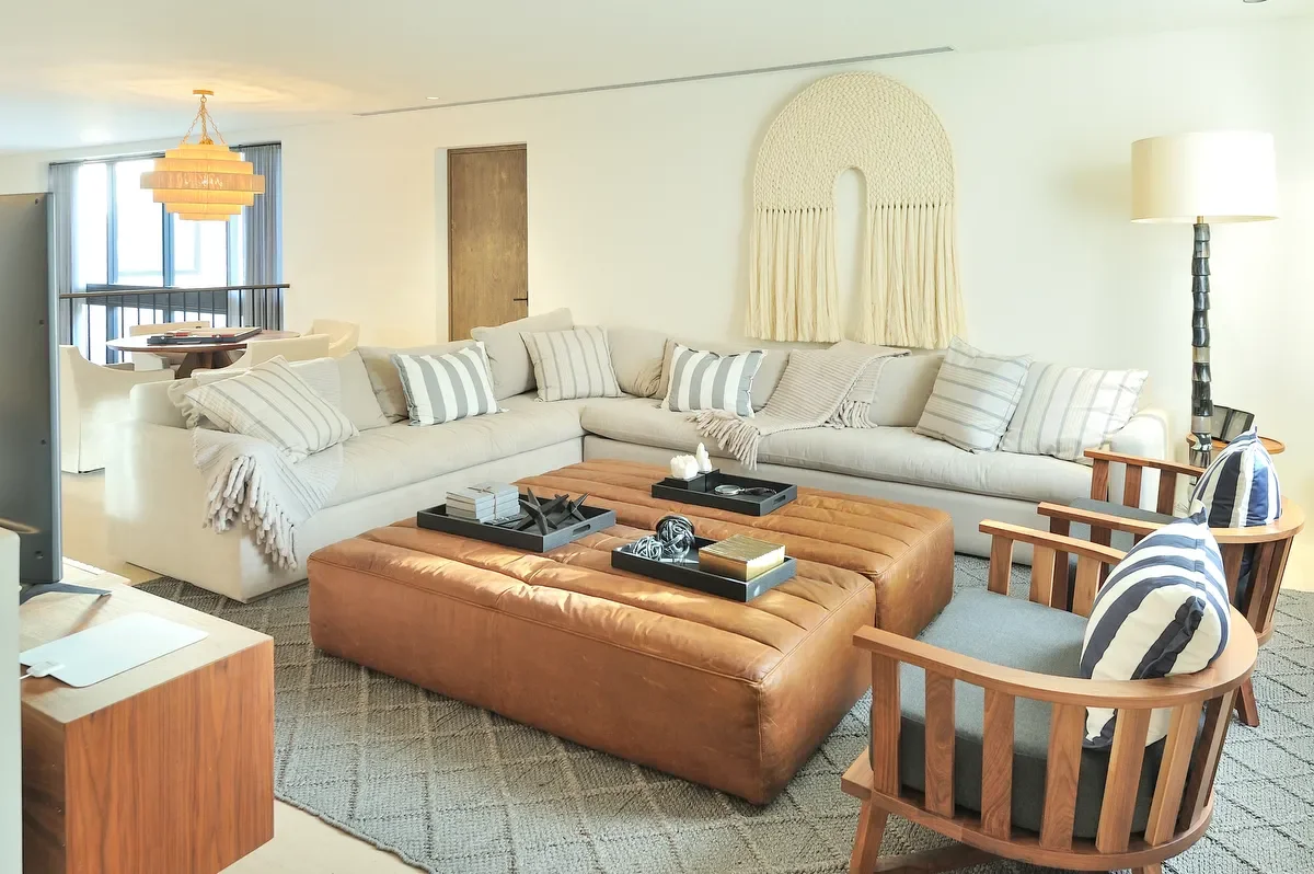 A modern living room with a large sectional sofa, striped pillows, two wooden armchairs with striped cushions, a leather ottoman, a side lamp, and a decorative wall hanging.