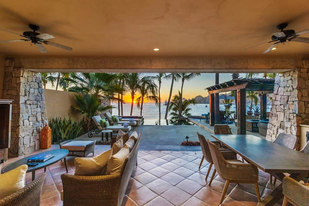 Indoor living space with stone walls, ceiling fans, and a large open window view of a tropical sunset over the ocean, with palm trees and outdoor furniture.