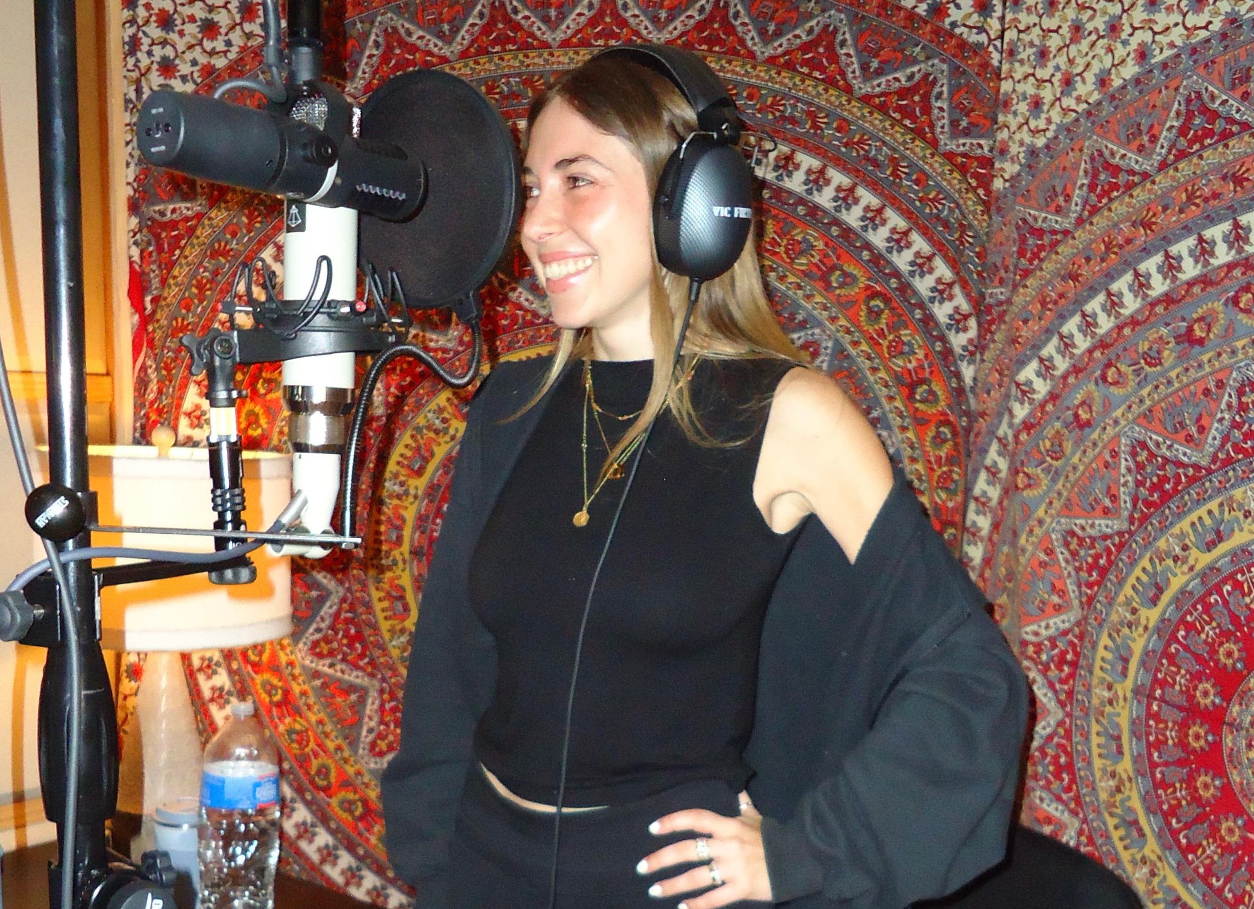 Young woman recording vocals in a music studio, wearing headphones, smiling, with a microphone in front of her and a colorful tapestry in the background.