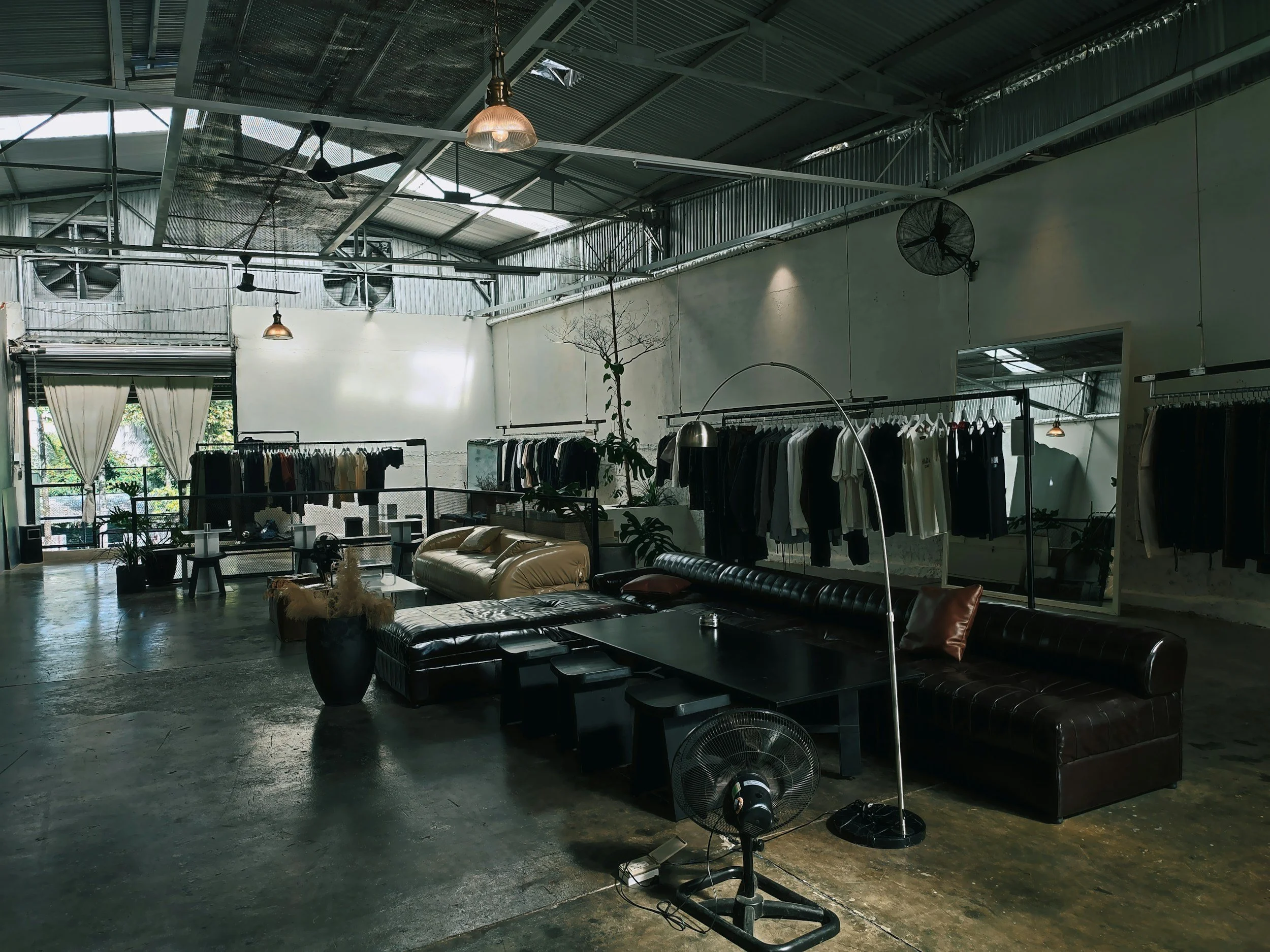 Interior of a clothing store with racks of clothes, a leather couch, potted plants, ceiling fans, and industrial-style metal ceiling.