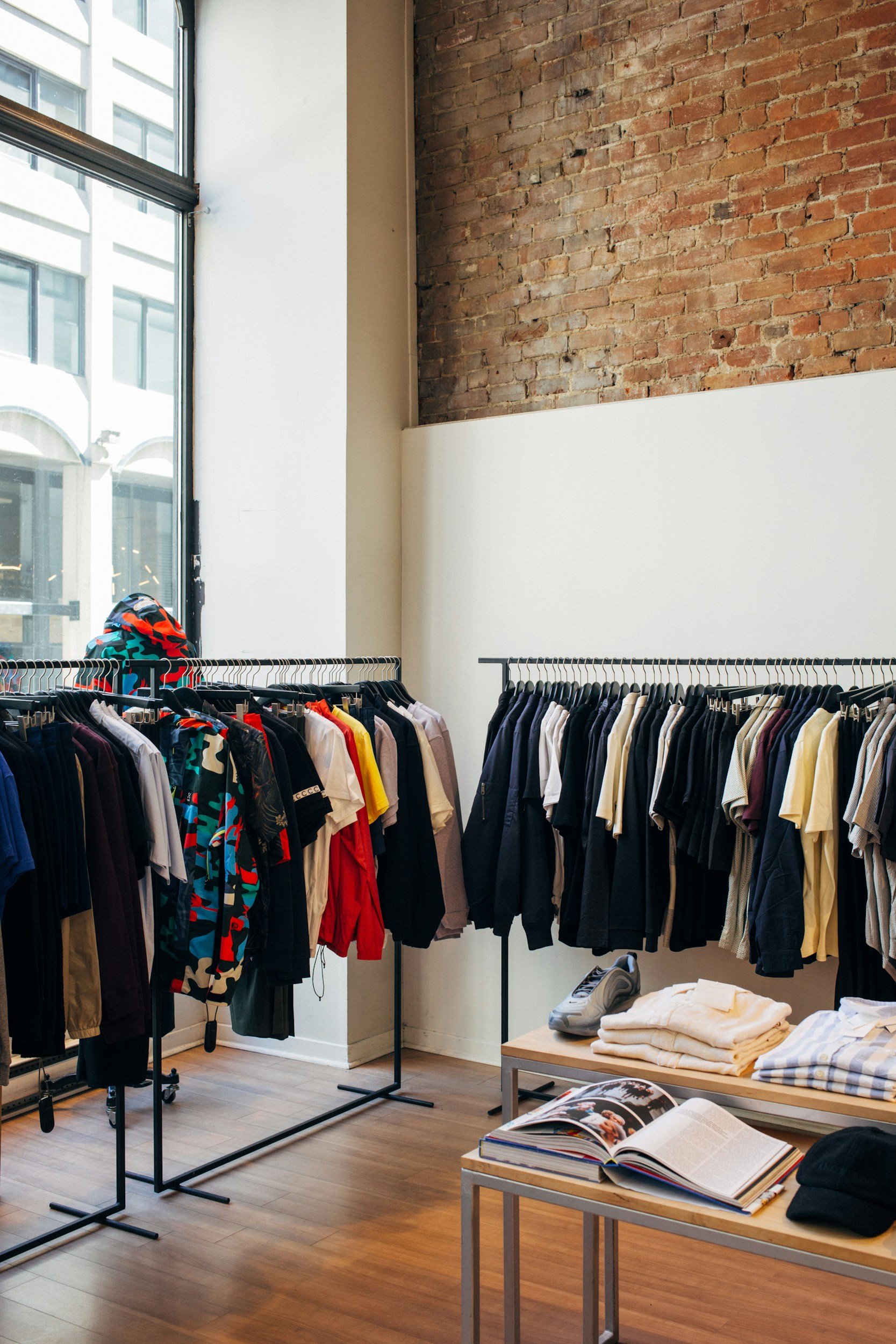 Inside a clothing store with racks of assorted clothes and a table with folded shirts and magazines, set against a brick wall and large windows.