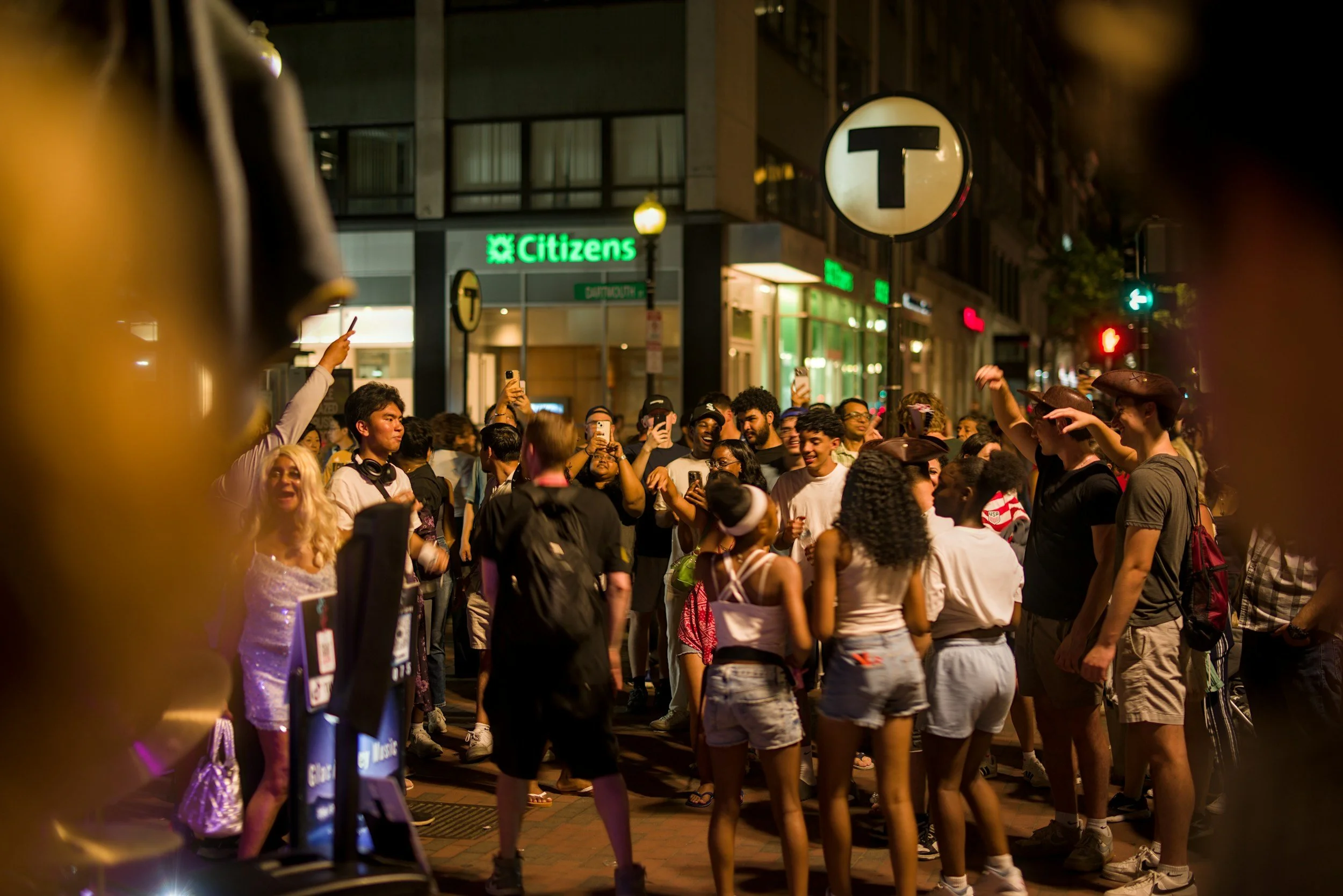 A crowd of people gathered on a city street at night, some taking photos and videos. There are illuminated store signs, including a green 'Citizens' sign. The street has a large round sign with a letter 'T', indicating a transit station entrance.
