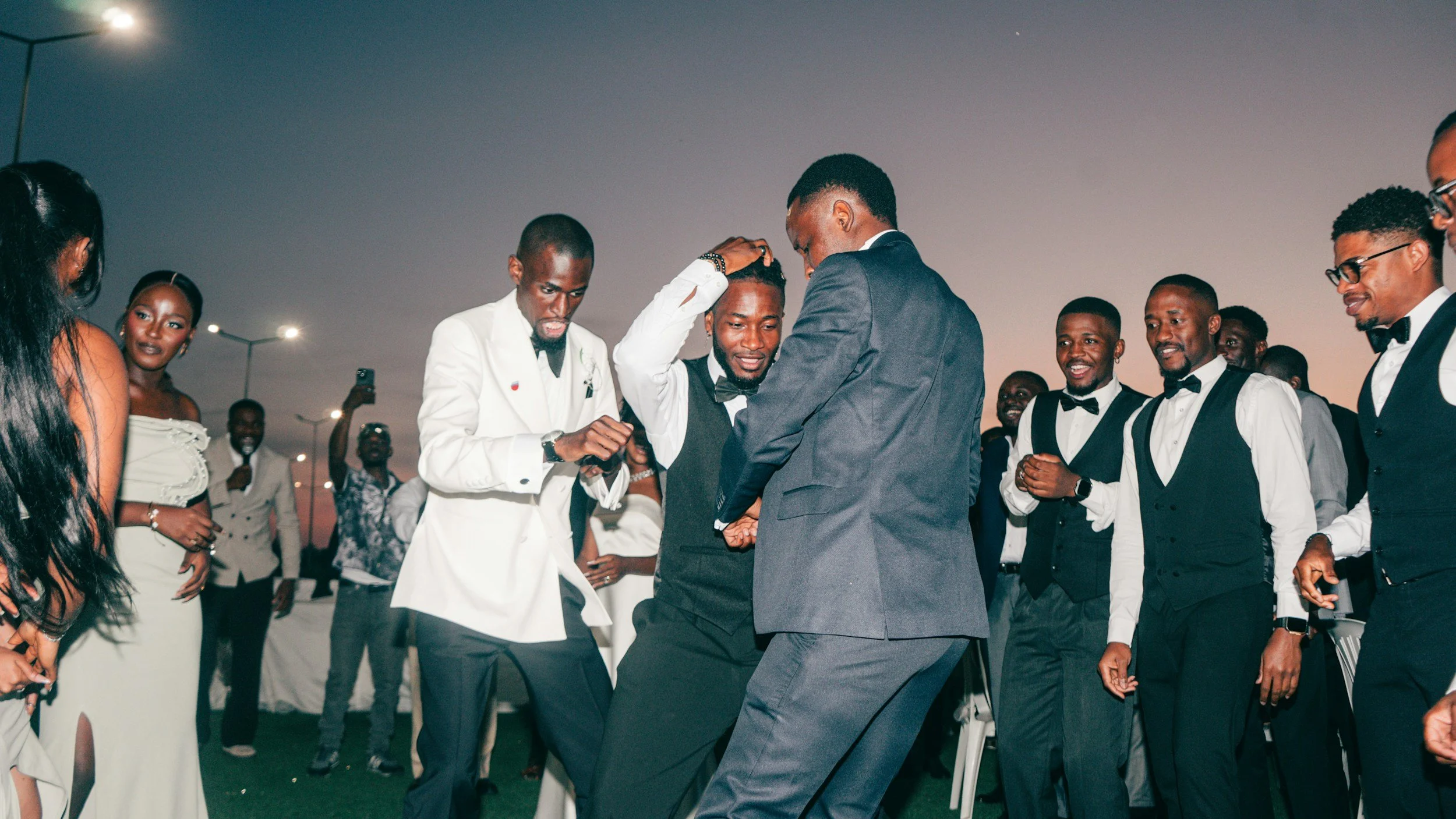 A group of well-dressed people dancing and smiling at an outdoor event during sunset, with some individuals wearing tuxedos and others in elegant dresses.