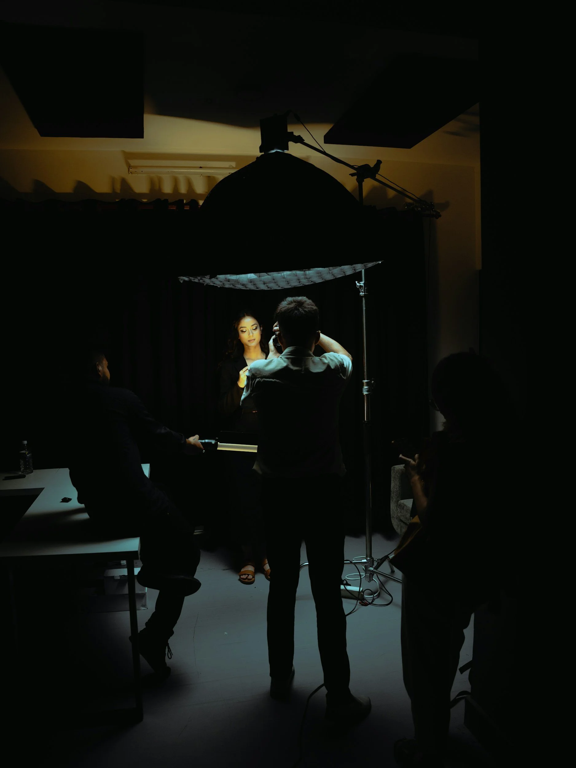 Scene inside a photography studio with a model standing under a large umbrella reflector, and several people adjusting equipment or taking photos, with dark surroundings and bright studio lighting.