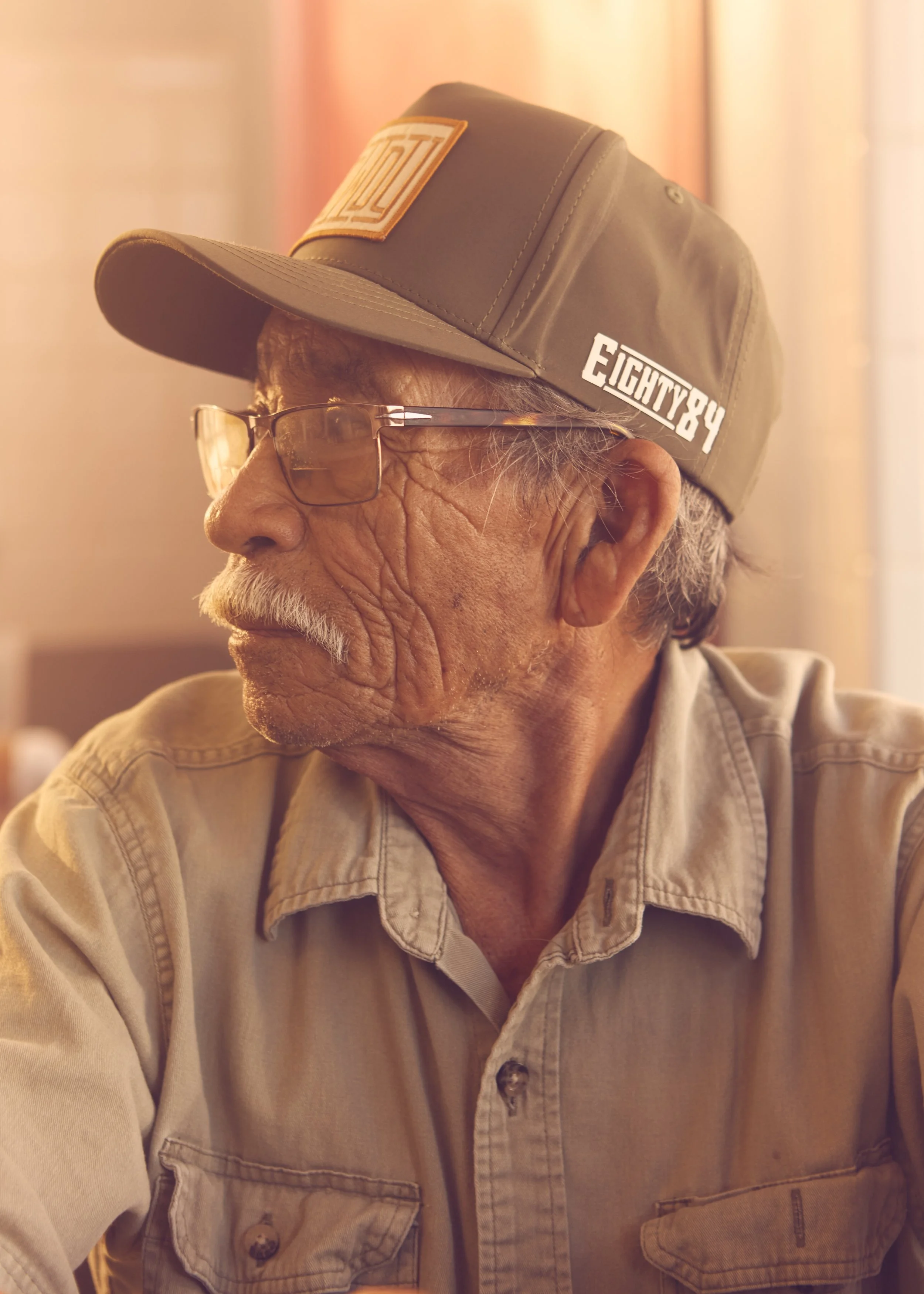 A close-up side profile of an elderly man wearing glasses, a beige button-up shirt, and a gray baseball cap with patches that read 'FIGHT86' and some other text, in an indoor setting with warm lighting.