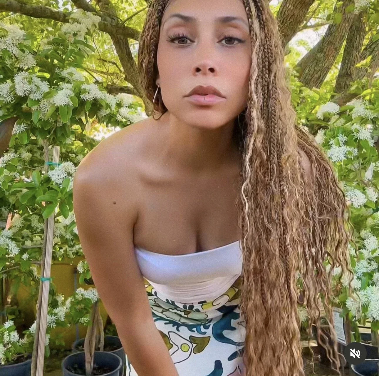 A woman with long, wavy, light brown hair poses outdoors in front of green leafy trees and white flowering plants.