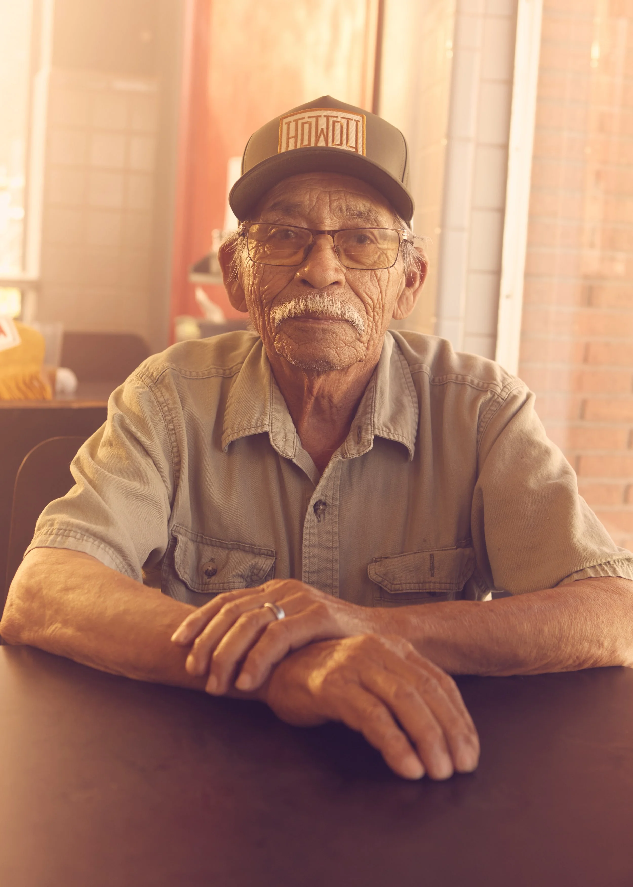 An elderly man with white hair, glasses, and a mustache, wearing a beige shirt and a baseball cap with 'HOU'DU' on it, sitting indoors near a brick wall, resting his arms on a table with hands clasped.