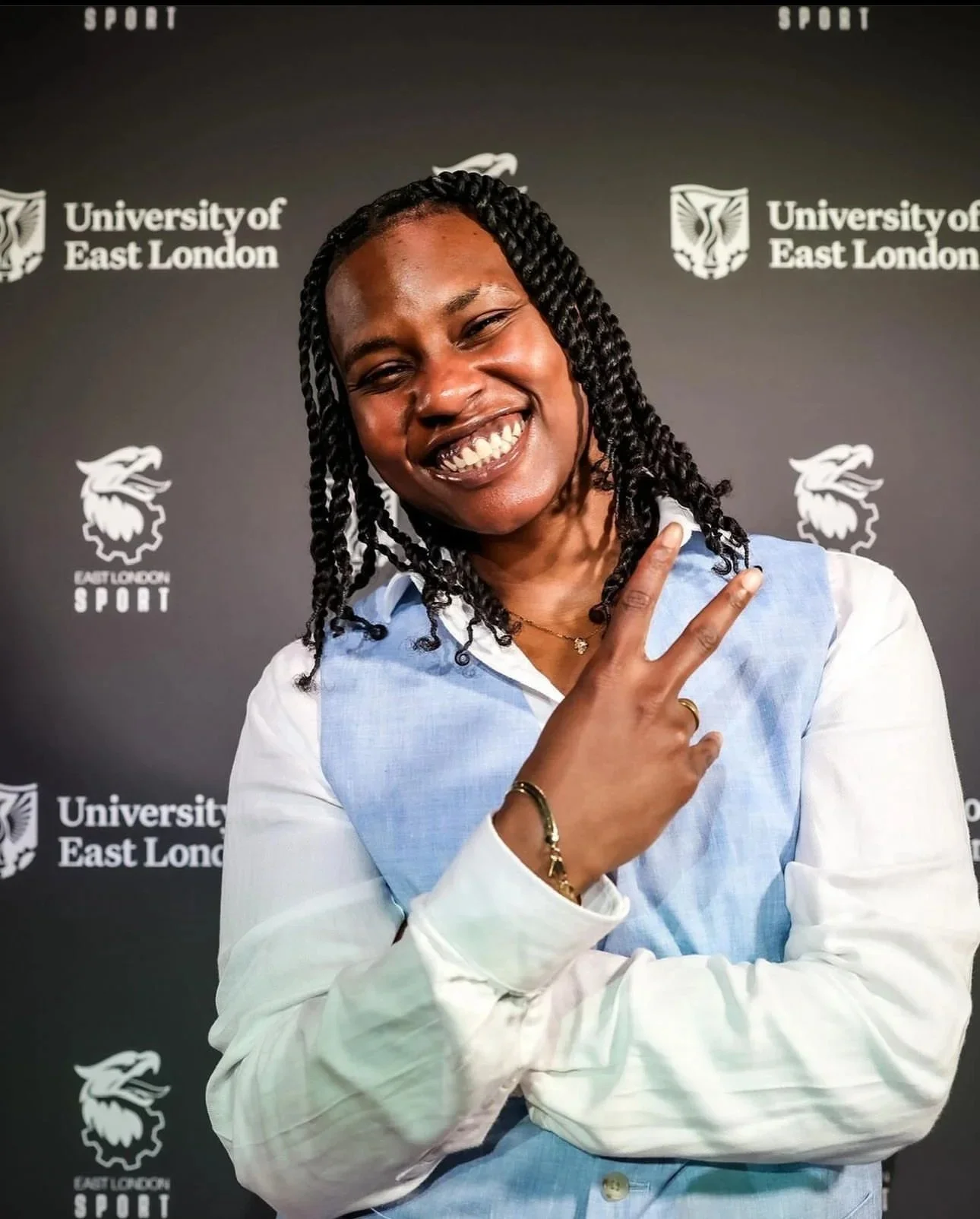 A young woman in a light blue vest and white shirt smiling and making a peace sign with her right hand, standing in front of a black background with the logos of the University of East London and East London Sport.