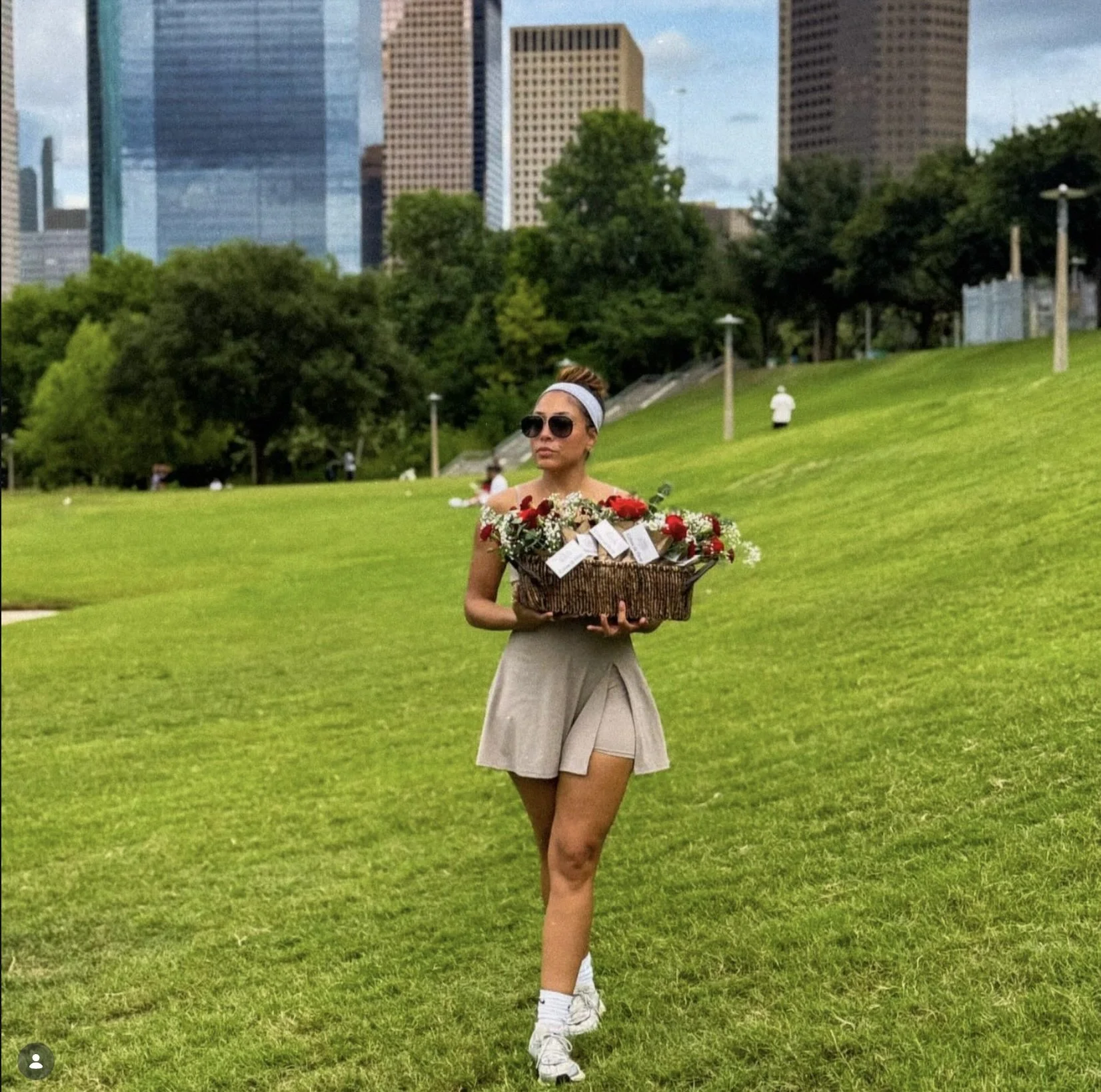 A woman in a beige skirt, white sneakers, sunglasses, and a gray headband is walking on green grass in an urban park, holding a large basket of flowers.