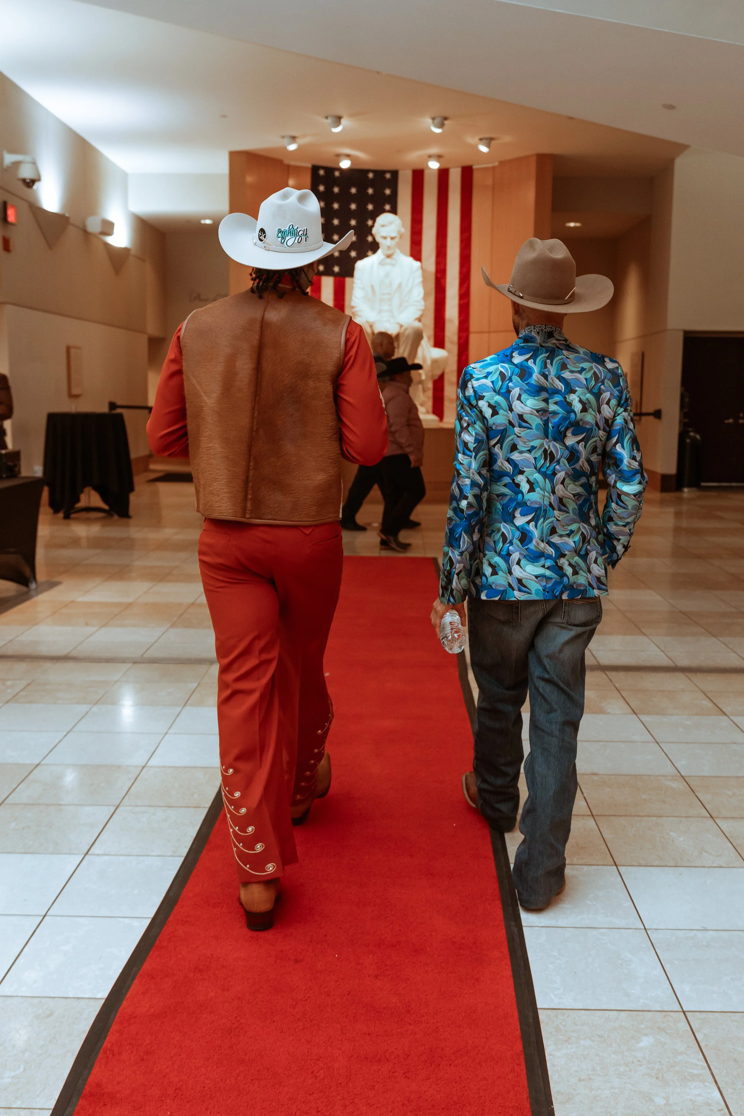 Two people walking on a red carpet towards a white statue with a U.S. flag in the background. The person on the left wears a cowboy hat, a brown vest, and red pants. The person on the right wears a beige cowboy hat, a floral shirt, and jeans, and is holding a plastic water bottle.